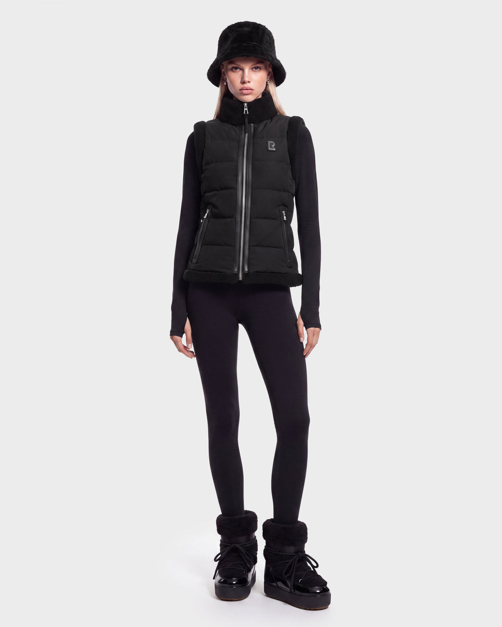 A woman stands facing forward against a plain white background, wearing the Rudsak HALI - 8126771 Black puffer vest filled with RDS-certified down for lightweight warmth, paired with a black bucket hat, long-sleeve black top, black leggings, and black boots.