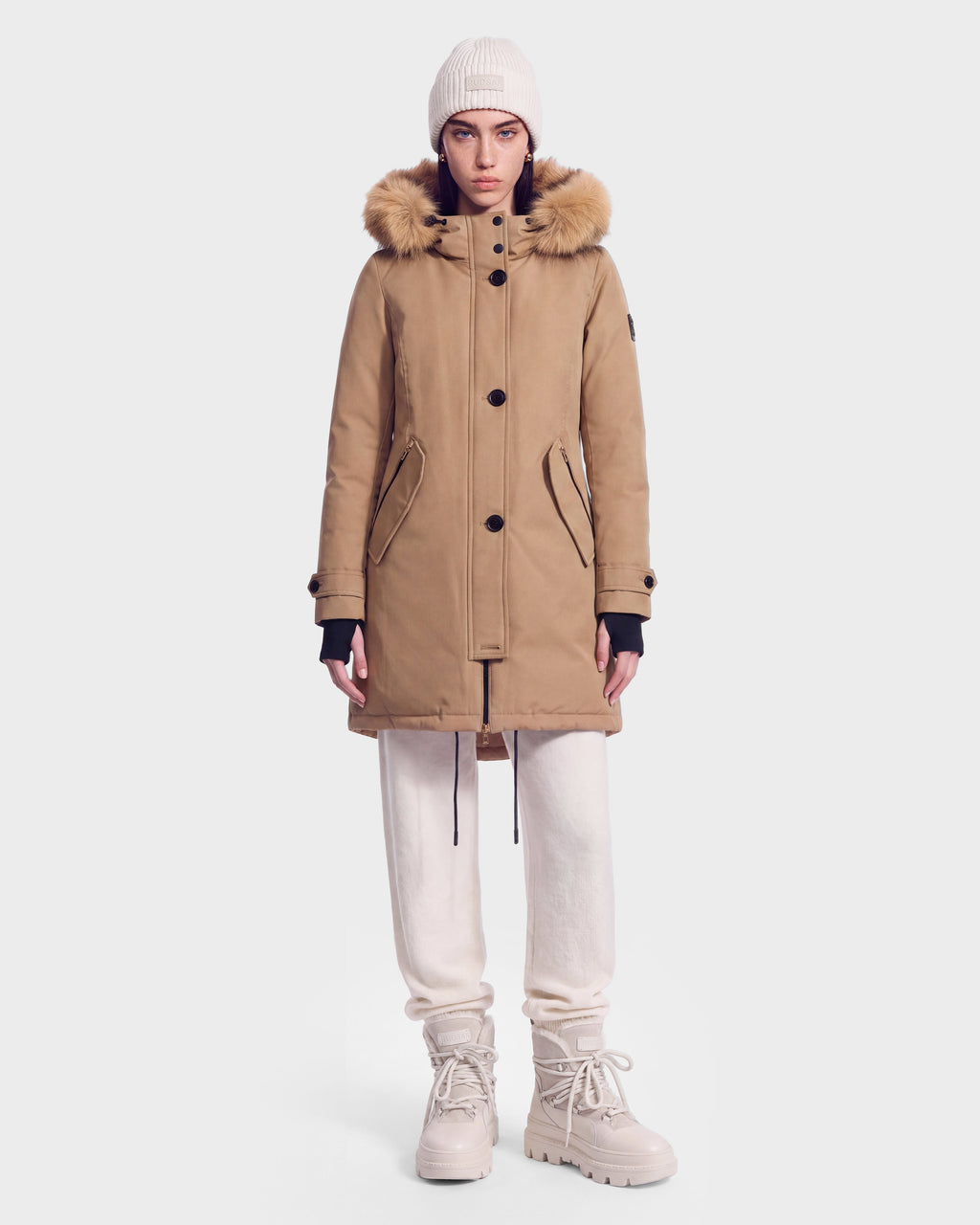 A person wearing the Rudsak KEYSHIA - 8126517 Camel women’s down parka, which features a fur-lined hood and is filled with RDS-certified down for guaranteed warmth and comfort, stands facing forward against a plain white background. She pairs the winter coat with a white beanie, white pants, and beige boots.