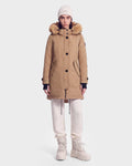 A person wearing the Rudsak KEYSHIA - 8126517 Camel women’s down parka, which features a fur-lined hood and is filled with RDS-certified down for guaranteed warmth and comfort, stands facing forward against a plain white background. She pairs the winter coat with a white beanie, white pants, and beige boots.