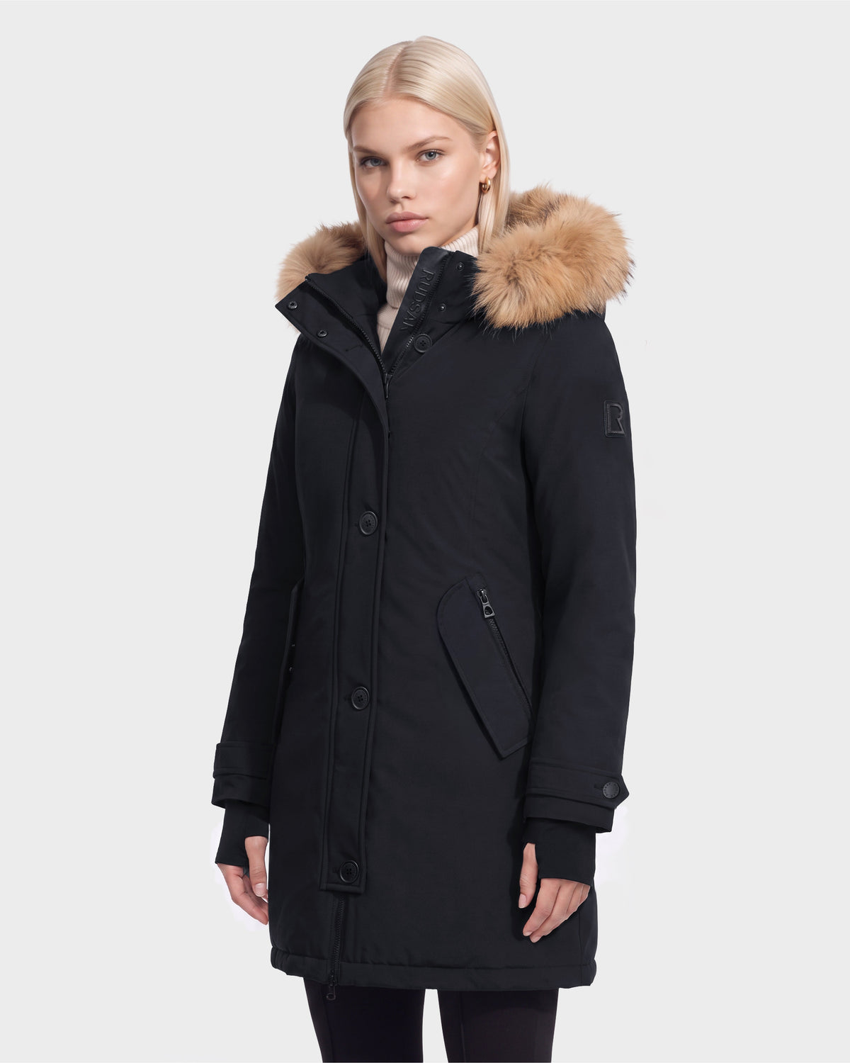 A woman with straight blonde hair wears the Rudsak KEYSHIA - 8126517 Black women’s down parka with fur-lined hood and angled zippered pocket, featuring RDS-certified down, against a plain white background.