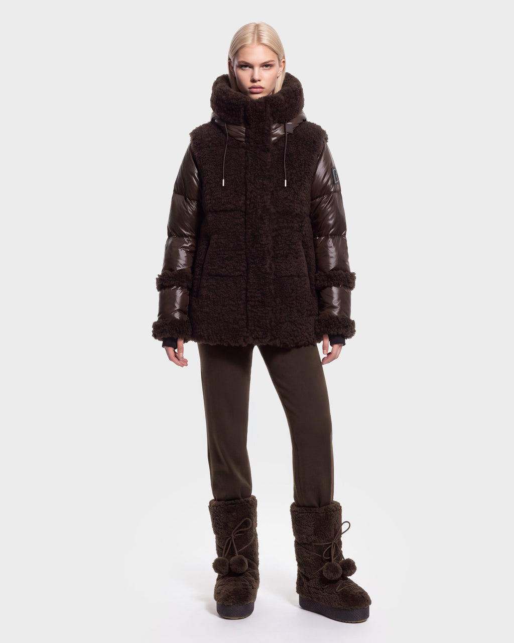 A person with straight blonde hair models the Rudsak MARISSA - 8126728 Espresso, a down-insulated winter coat in a dark, textured finish, styled with matching pants and furry boots with pom-poms, standing against a plain light background.