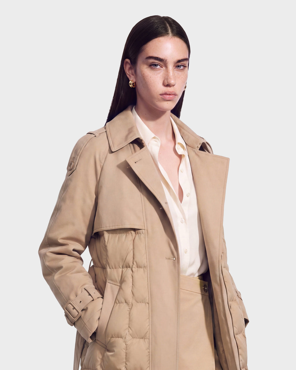A woman with straight brown hair wears the Rudsak ELVIE - 8126512 Almond beige down trench coat over a cream blouse and matching pants. She stands against a plain light background, looking forward with a neutral expression.