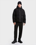 A person stands facing forward against a plain white background, wearing the Rudsak STERLING EL - 6126759 Black puffer jacket, paired with a black beanie, black pants, and black sneakers.