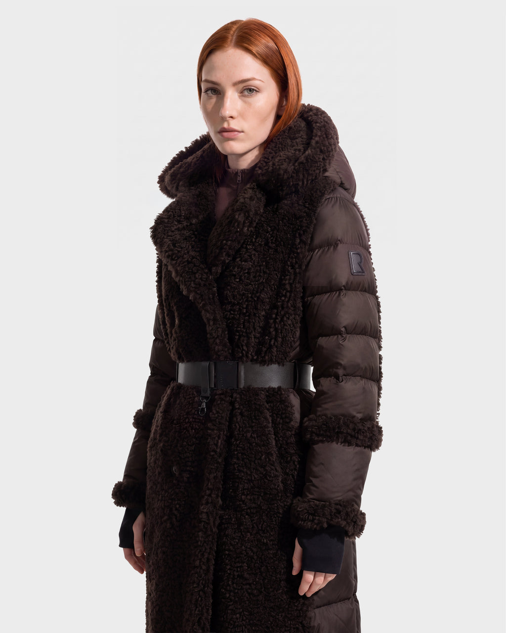 A person with red hair wears the Rudsak NOELLE - 8126971 Espresso, a long, dark brown (espresso) hooded winter coat featuring a thick collar, black belt, and logo patch on the sleeve, standing against a plain white background.