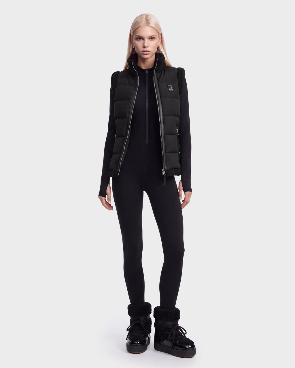 A person with long blonde hair wears the Rudsak HALI - 8126771 Black puffer vest filled with RDS-certified down, styled with a black fitted jumpsuit and black winter boots, standing against a plain white background.
