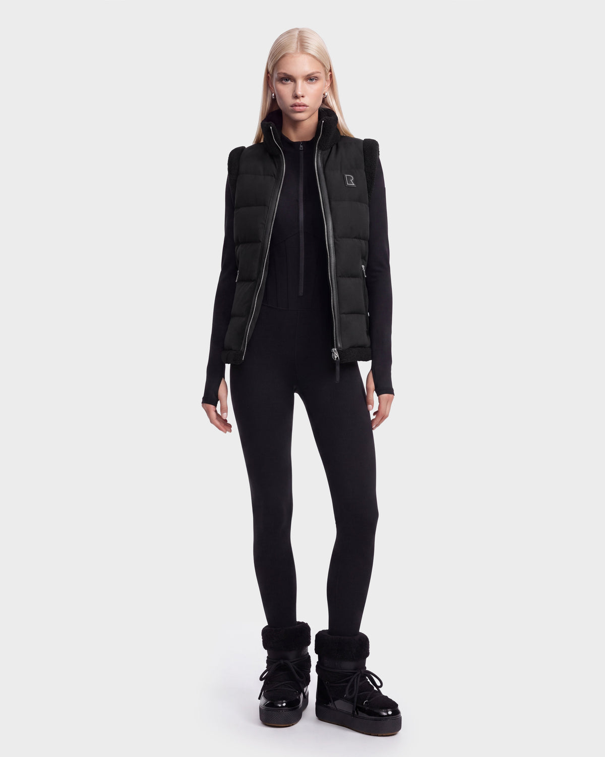 A person with long blonde hair wears the Rudsak HALI - 8126771 Black puffer vest filled with RDS-certified down, styled with a black fitted jumpsuit and black winter boots, standing against a plain white background.
