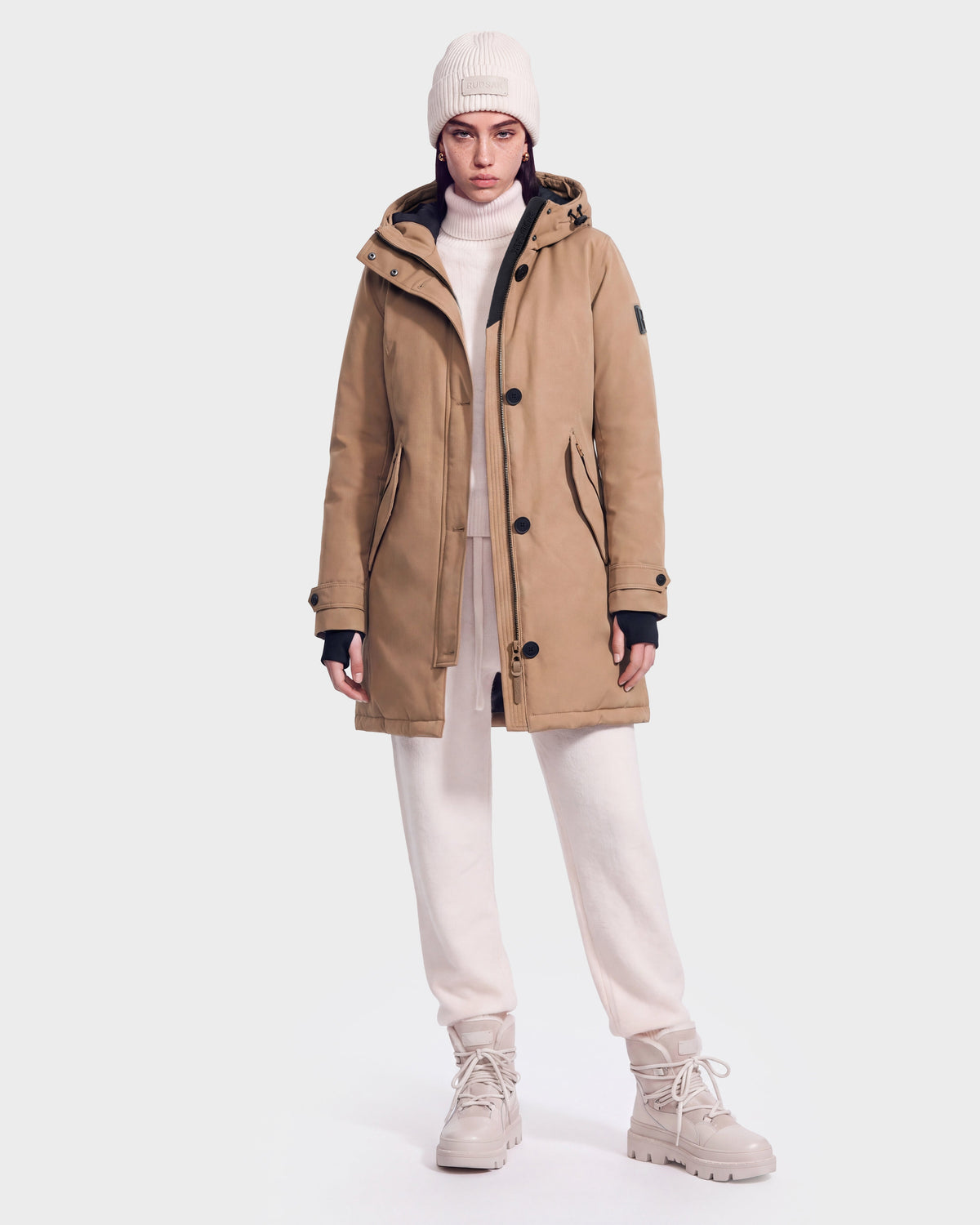 A person is wearing the Rudsak KEYSHIA - 8126517 Camel women’s down parka with RDS-certified down, styled with a white sweater, white pants, beige boots, and a white knit beanie. They are standing against a plain white background and looking straight at the camera with a neutral expression.