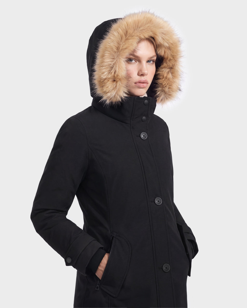 A person wearing the Rudsak KEYSHIA - 8126517 Black women’s parka with a fur-lined hood stands with hands in pockets, facing forward against a plain light background.