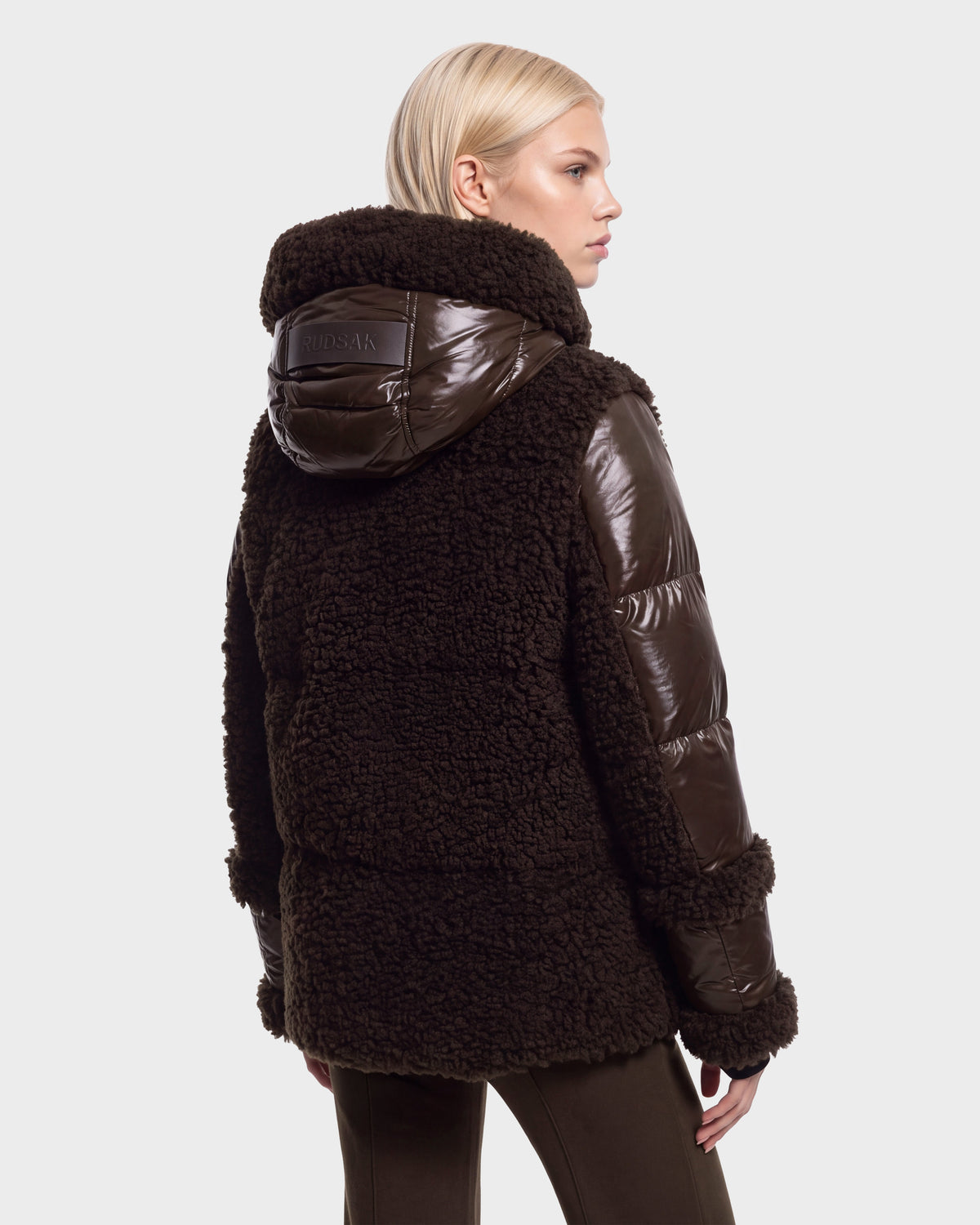 A person with short blonde hair is seen from the back against a plain white background, wearing the Rudsak MARISSA - 8126728 Espresso: a dark brown, hooded shearling and puffer jacket—perfect winter outerwear.