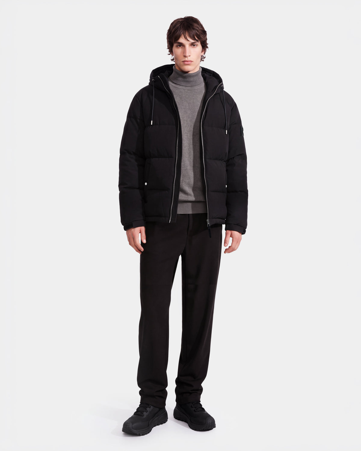 A person wearing the Rudsak STERLING EL - 6126759 Black puffer jacket over a gray turtleneck sweater and black pants stands against a plain white background.