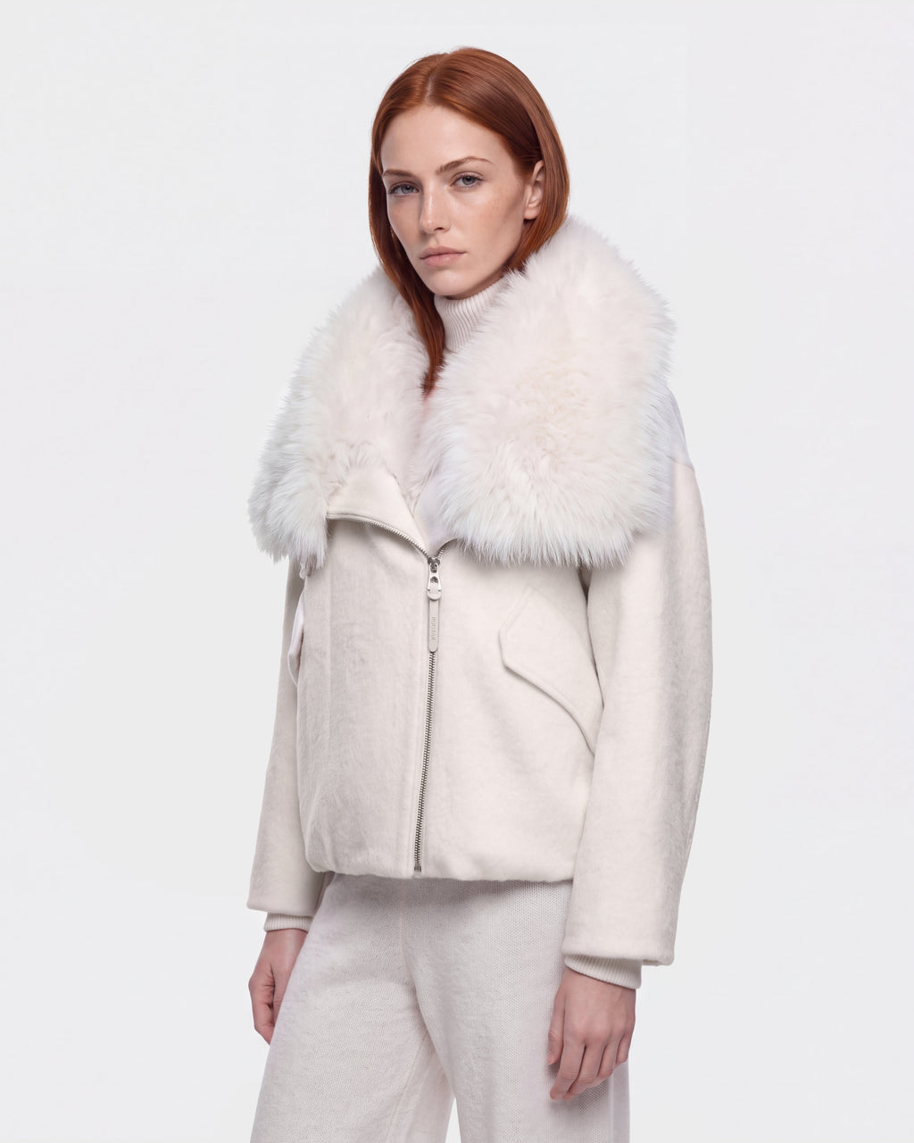 A woman with long red hair wears the Rudsak ZARIA - 8126999 Vanilla white winter puffer jacket, featuring a large, fluffy faux-fur collar, paired with white pants as she stands against a plain white background.
