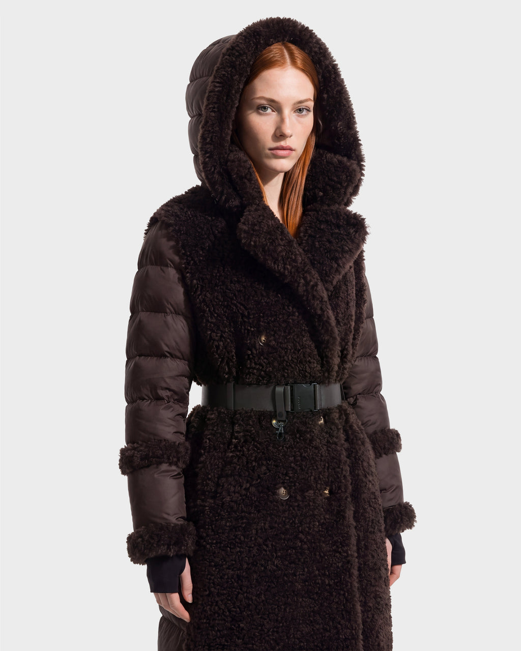 A woman with straight red hair wears the Rudsak NOELLE - 8126971 Espresso, a dark brown hooded winter coat with a textured faux fur front and sleeves, belted waist, and snap buttons. She faces forward against a plain, light background.