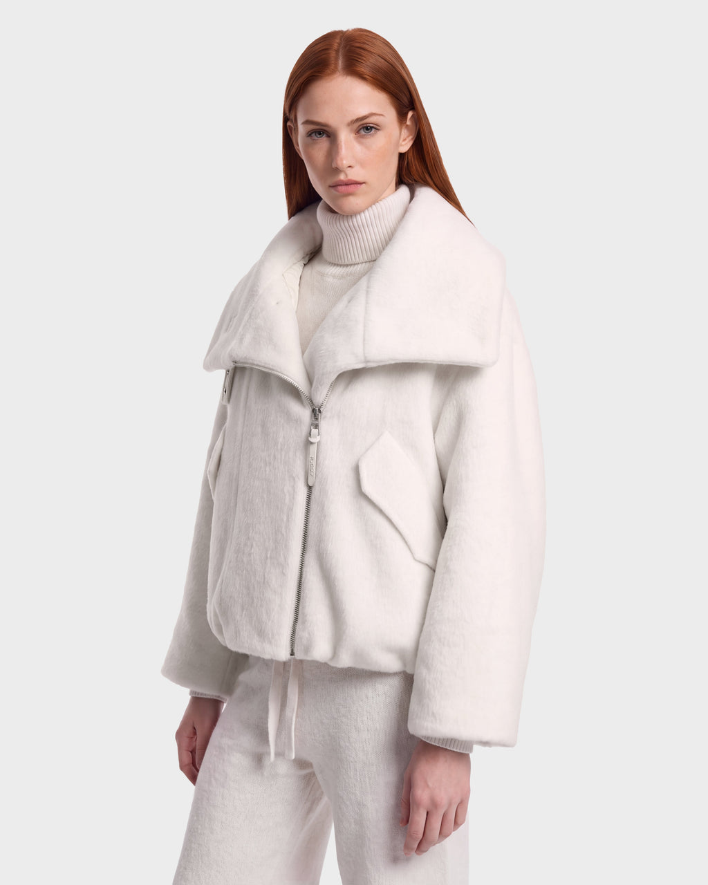 A woman with long red hair wears the Rudsak ZARIA - 8126999 Vanilla, a white oversized zip-up jacket with a large faux-fur collar and pockets, styled over a white turtleneck sweater and white pants, standing against a plain light gray background.