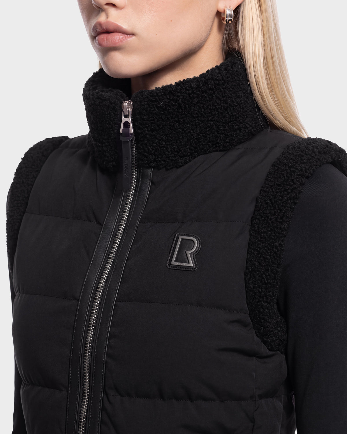 A woman with straight blonde hair wears the Rudsak HALI - 8126771 puffer vest in black, featuring a textured collar and shoulders, zip-up front, and a raised R logo on the chest. Designed for lightweight warmth, only her lower face and upper torso are visible.