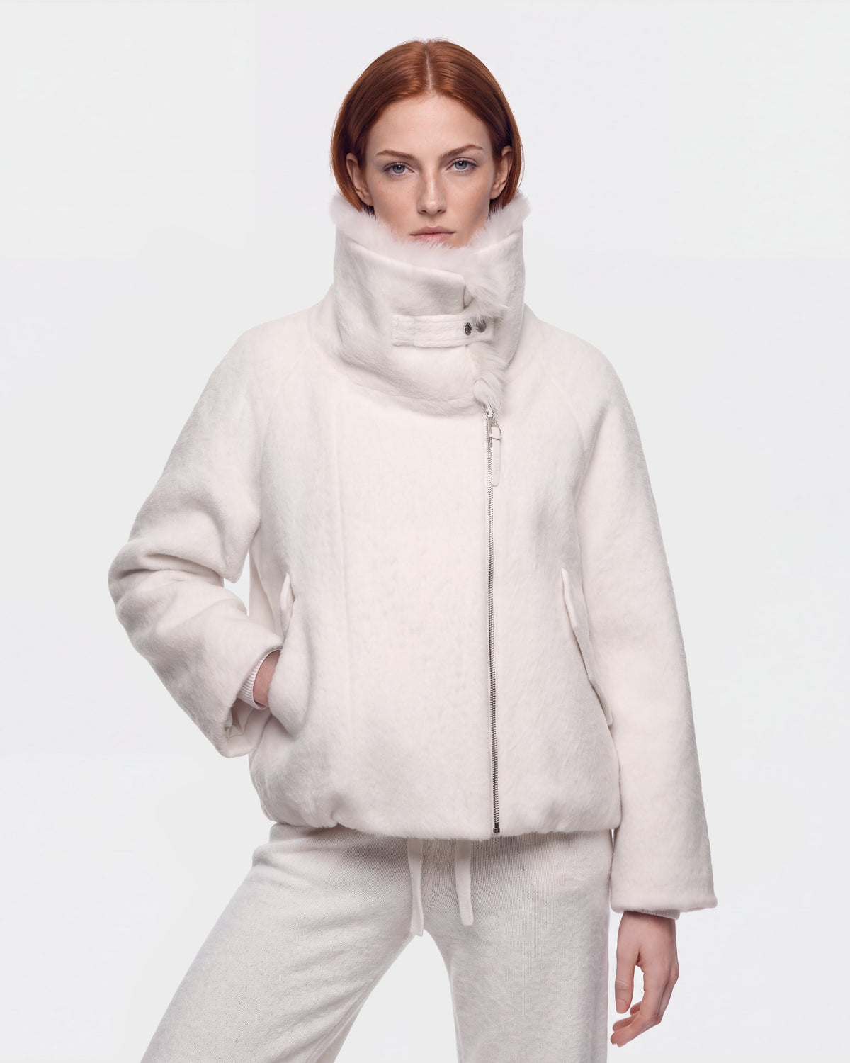 A woman with red hair stands against a plain white background, wearing the Rudsak ZARIA - 8126999 Vanilla high-collar winter puffer jacket with matching pants, one hand casually in her pocket.
