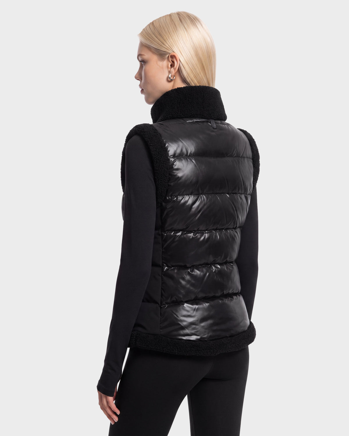 A person with straight blonde hair wears the Rudsak HALI - 8126771 Black, a shiny black puffer vest filled with RDS-certified down for lightweight warmth and accented with fuzzy trim, layered over a long-sleeve black top. They stand against a plain light background, facing away.