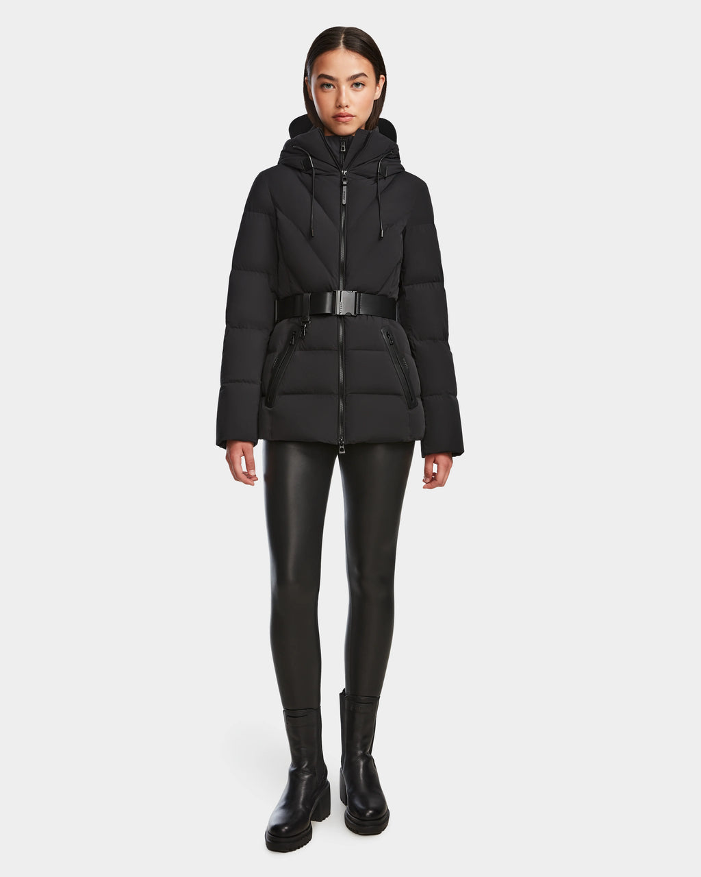A woman faces forward against a white background, wearing the Rudsak ENZAH - 8124692 Black mid-length puffer jacket with a belt, paired with black faux leather leggings and black ankle boots.