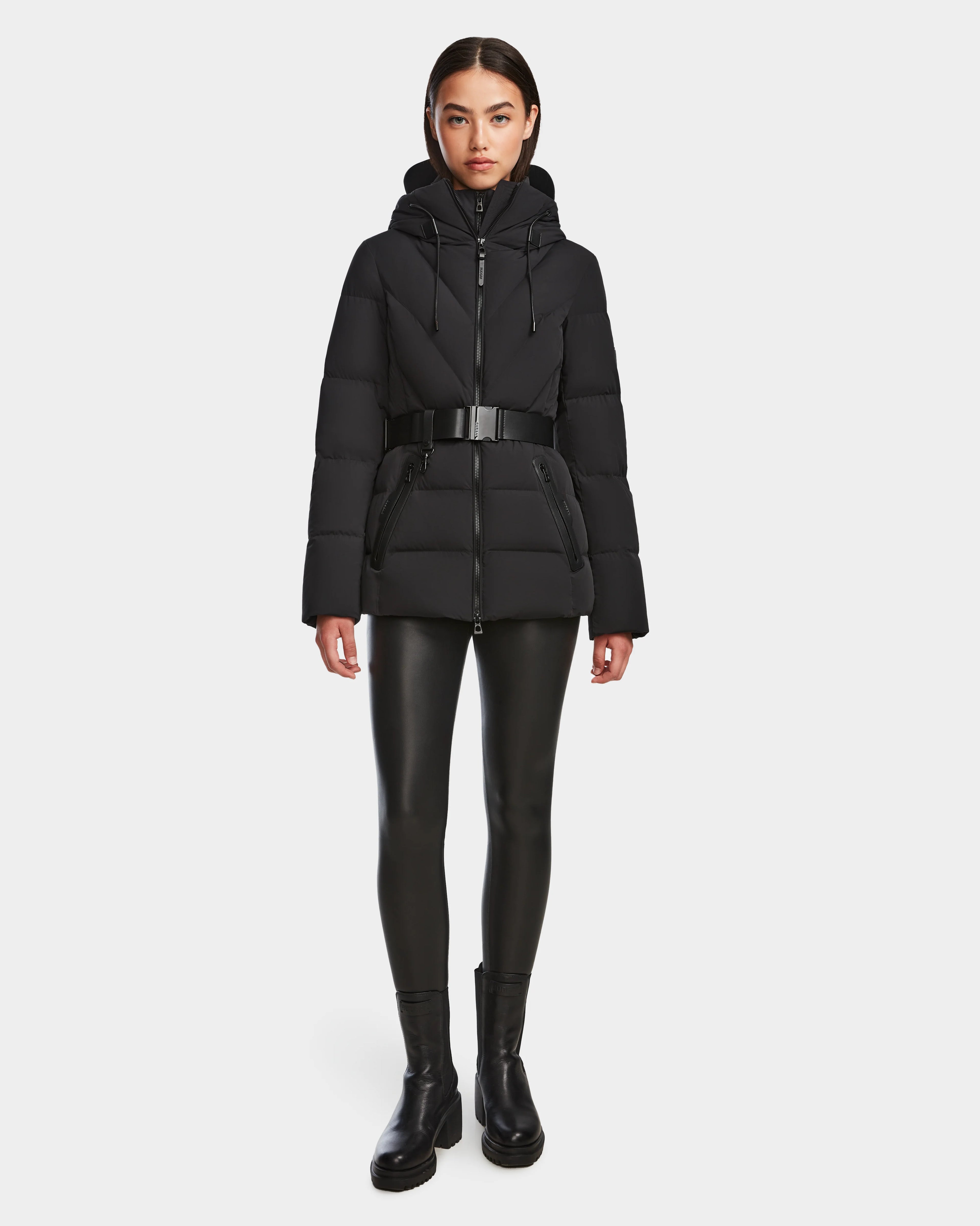 A woman faces forward against a white background, wearing the Rudsak ENZAH - 8124692 Black mid-length puffer jacket with a belt, paired with black faux leather leggings and black ankle boots.