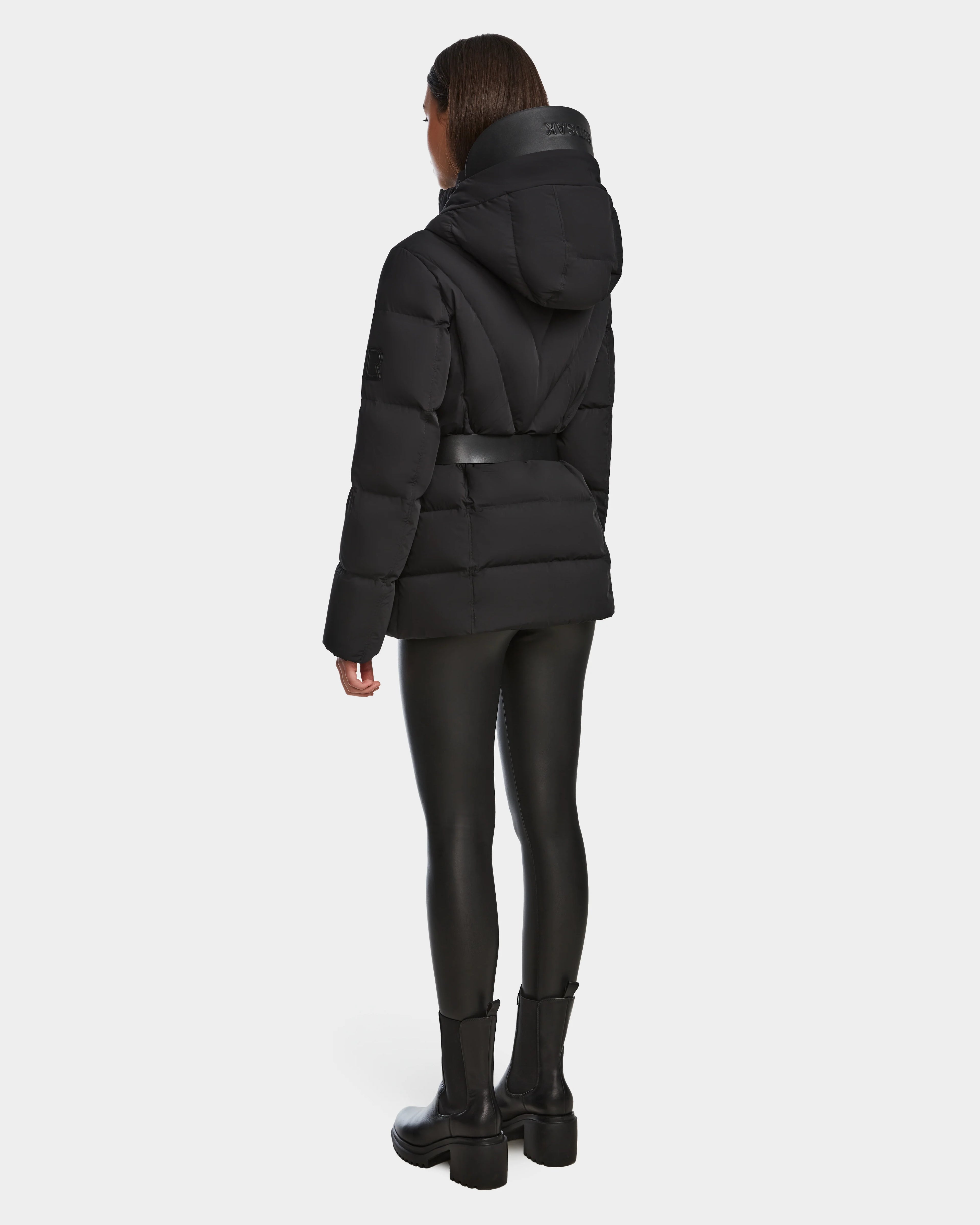 A person faces away in the Rudsak ENZAH - 8124692 Black womens down puffer jacket, belted at the waist with a hood, paired with black leggings and boots, set against a plain white background.