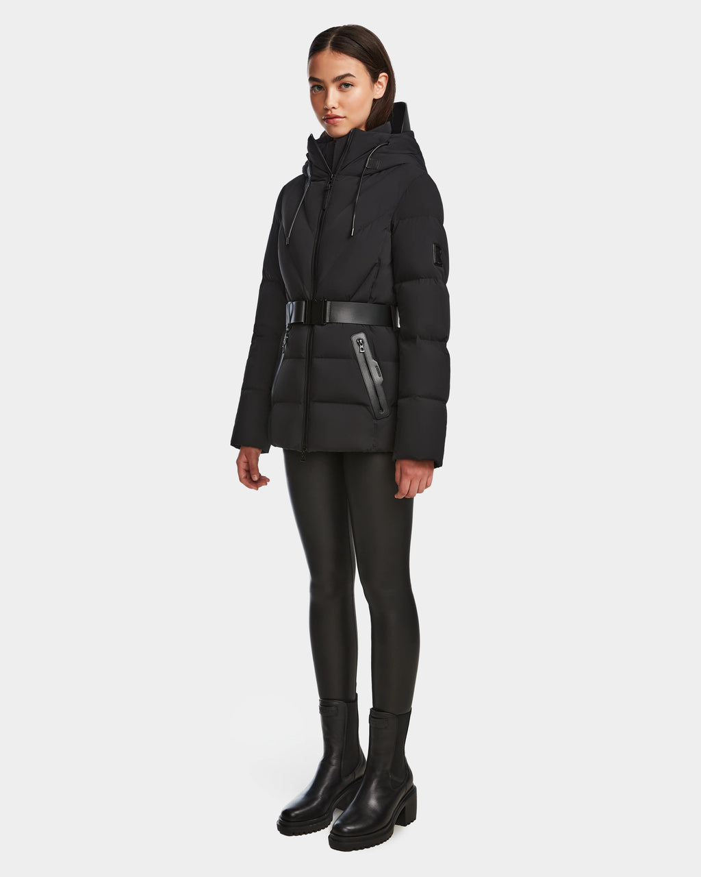 A woman stands slightly turned, wearing the Rudsak ENZAH - 8124692 Black mid-length puffer jacket with a belt, paired with black leggings and ankle boots, set against a plain white background.