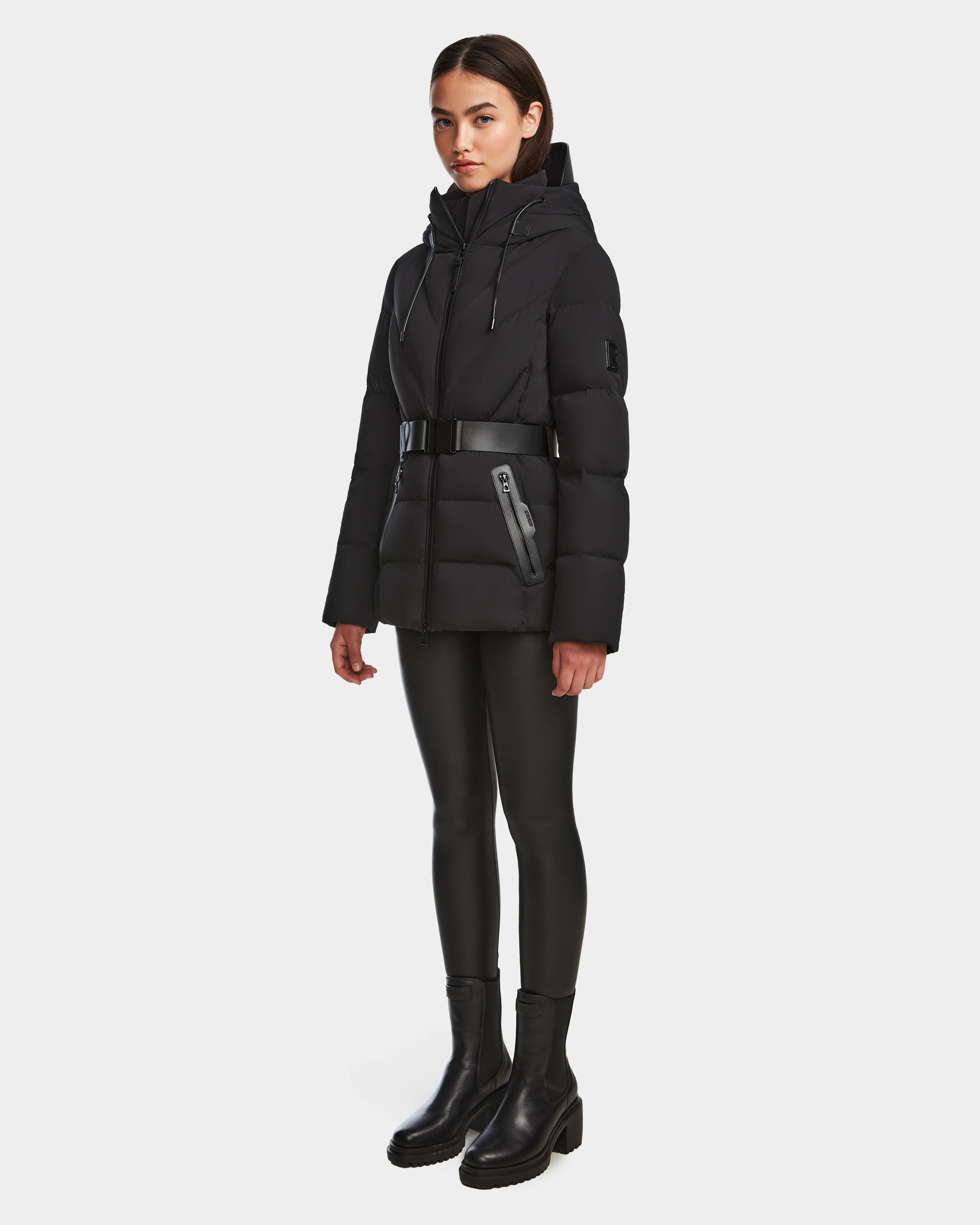 A woman stands slightly turned, wearing the Rudsak ENZAH - 8124692 Black mid-length puffer jacket with a belt, paired with black leggings and ankle boots, set against a plain white background.