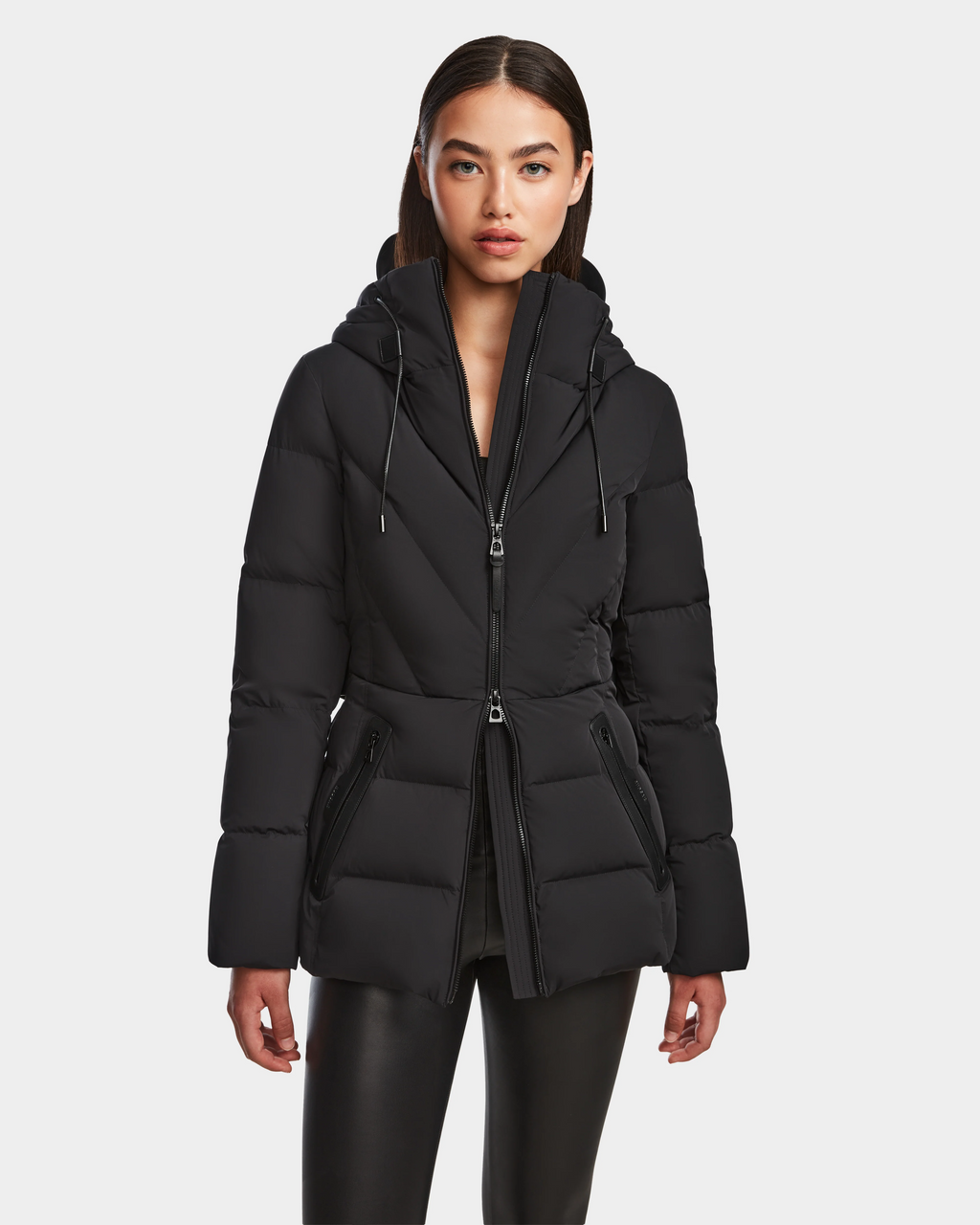 A woman with straight dark hair models the Rudsak ENZAH - 8124692 Black mid-length puffer jacket, featuring a hood and zippered pockets. She stands facing forward in black pants against a plain white background.