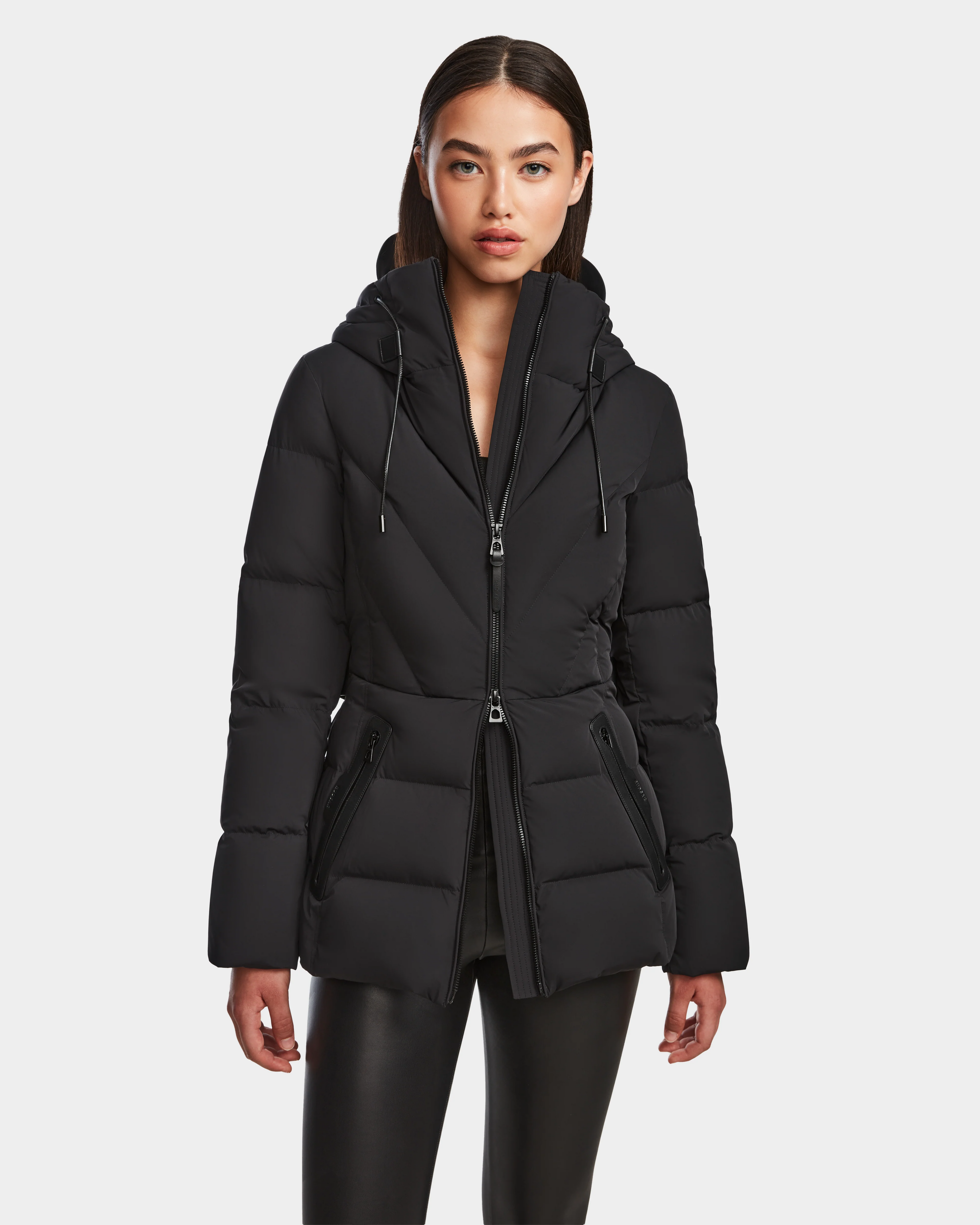 A woman with straight dark hair models the Rudsak ENZAH - 8124692 Black mid-length puffer jacket, featuring a hood and zippered pockets. She stands facing forward in black pants against a plain white background.