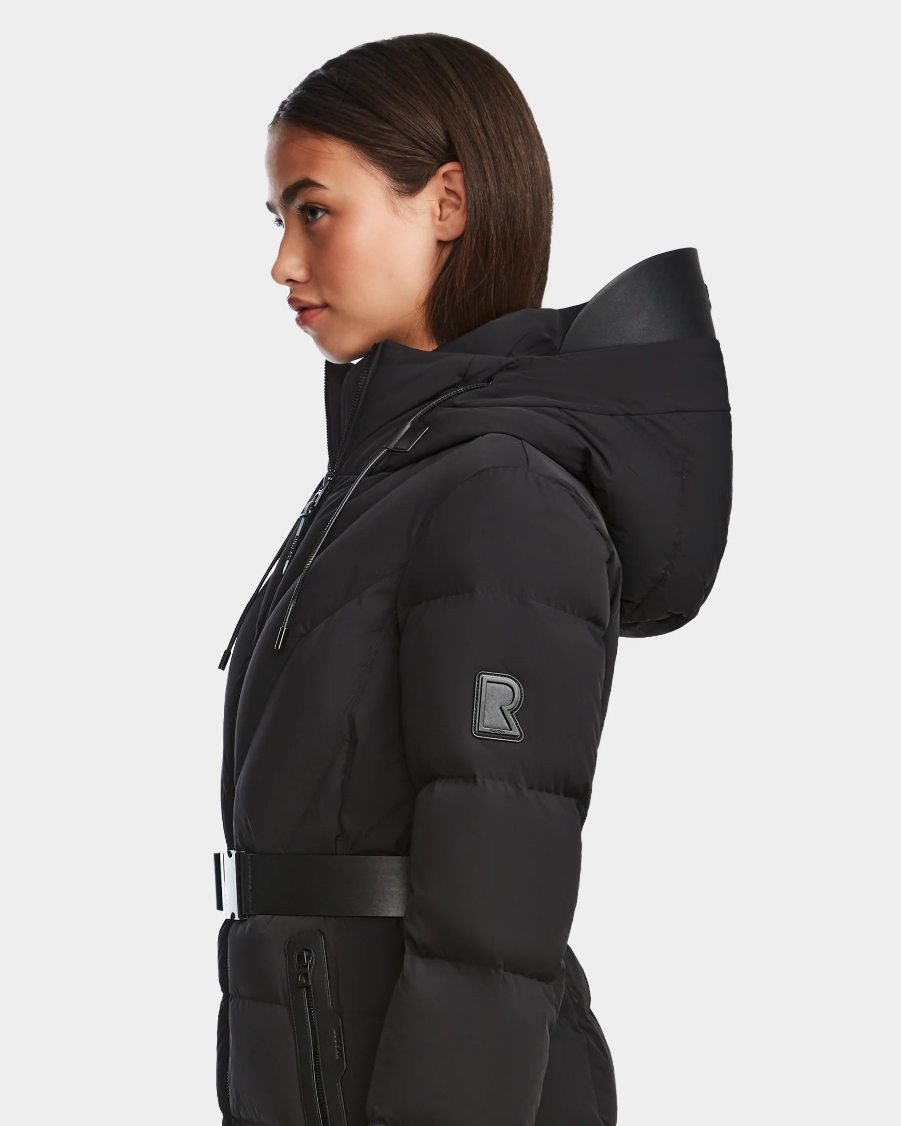 A woman in profile wears the Rudsak ENZAH - 8124692 Black mid-length puffer jacket with a hood, belted waist, and a large “R” patch on the sleeve, standing against a plain white background.