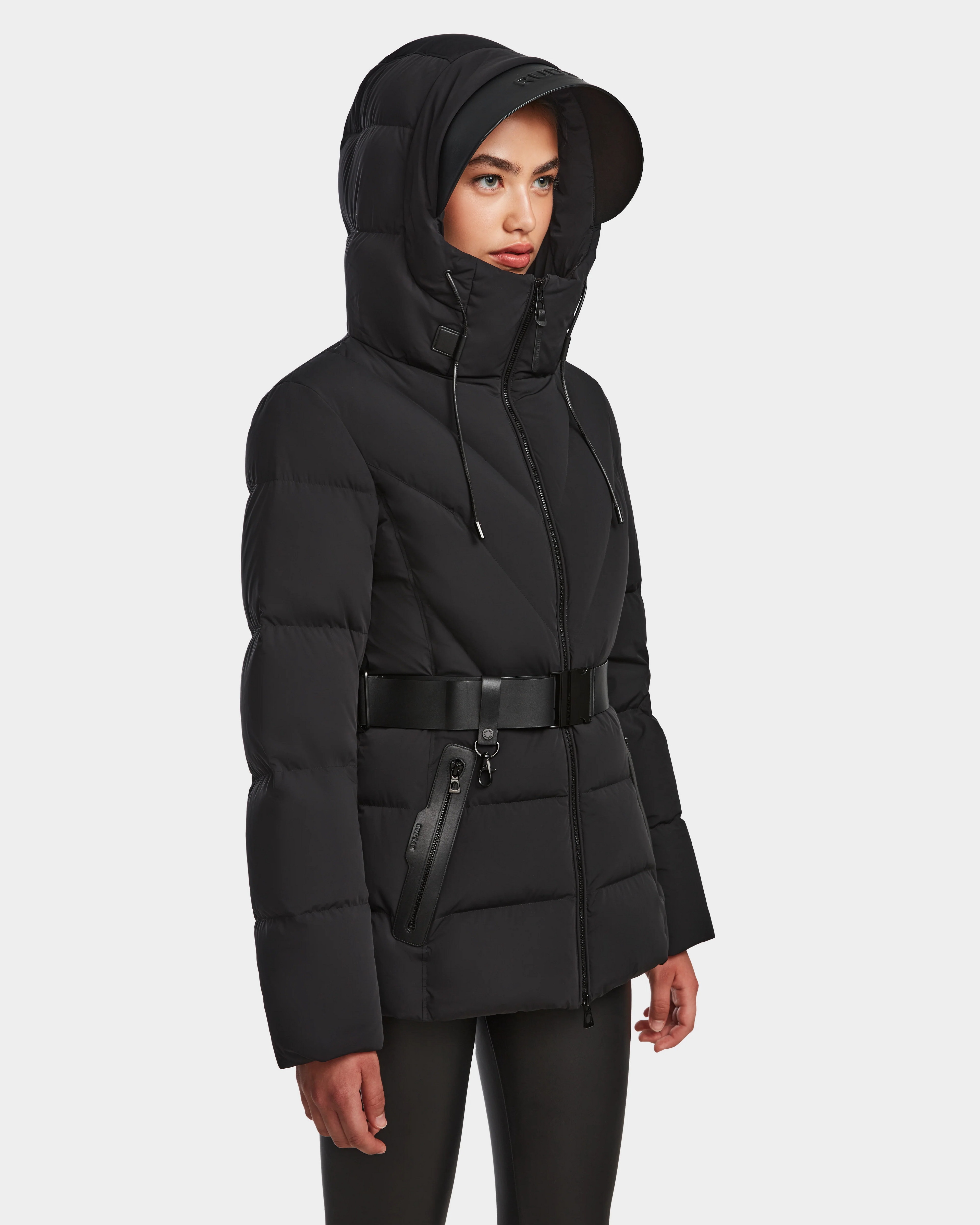 A woman models the Rudsak ENZAH - 8124692 Black, a mid-length womens down puffer jacket with hood, high collar, and wide belt—perfect for stylish winter looks—against a plain light background.