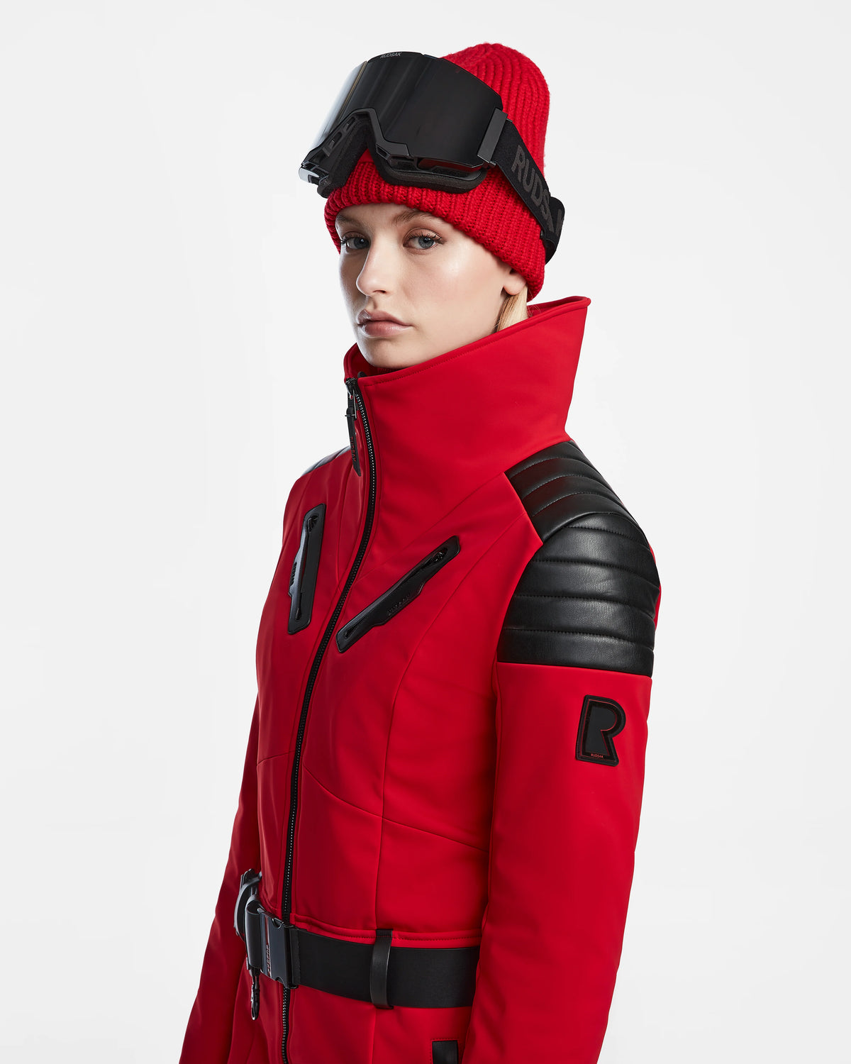 A person in the Rudsak ARLEY - 8124717 Flame Red ski jacket with black shoulder accents, a red knit beanie, and black ski goggles poses against a white backdrop, highlighting RECCO rescue technology.