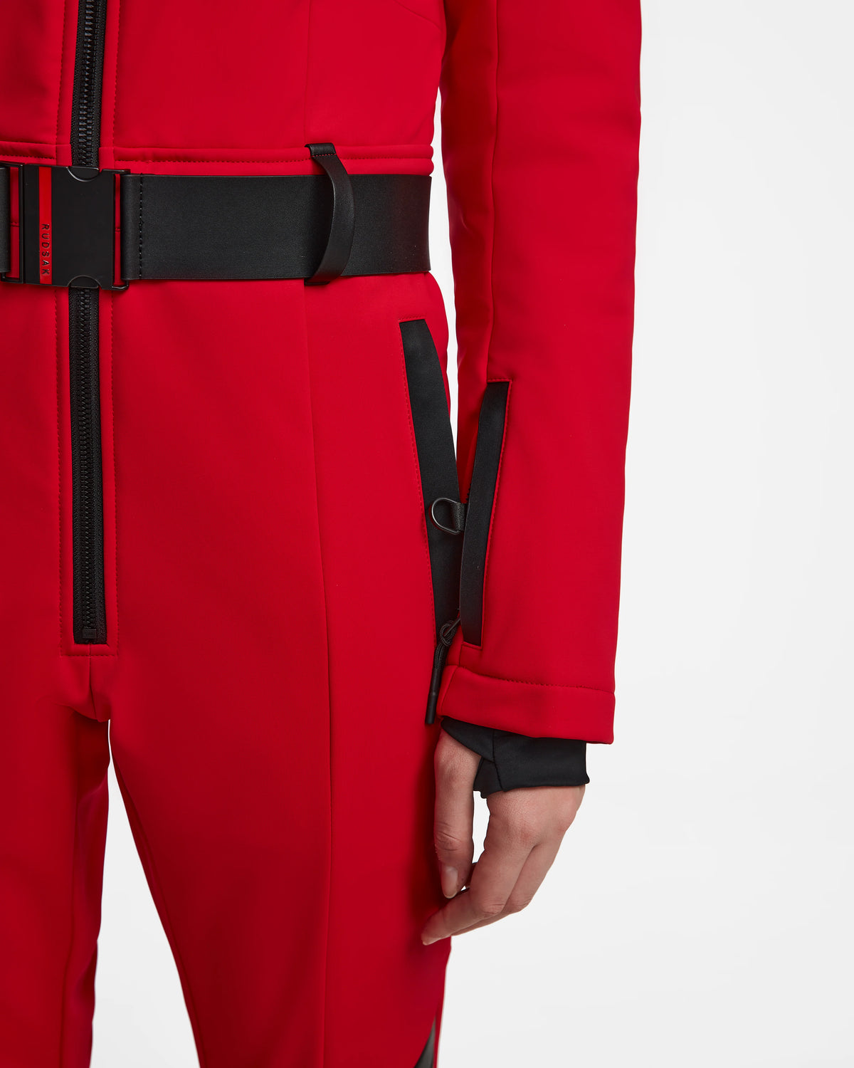 A person wears the Rudsak ARLEY - 8124717 Flame Red fitted women’s ski suit with black accents and a black belt, standing relaxed with a sleeve zippered pocket visible.