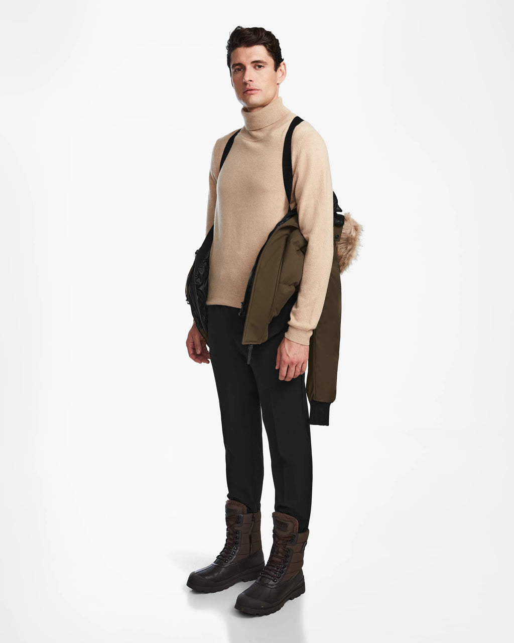 A man faces forward in a beige turtleneck, black pants, and brown boots, holding the Rudsak DALLAS - M122501 Army weather-resistant puffer jacket with a fur-lined hood over his shoulder against a white background.