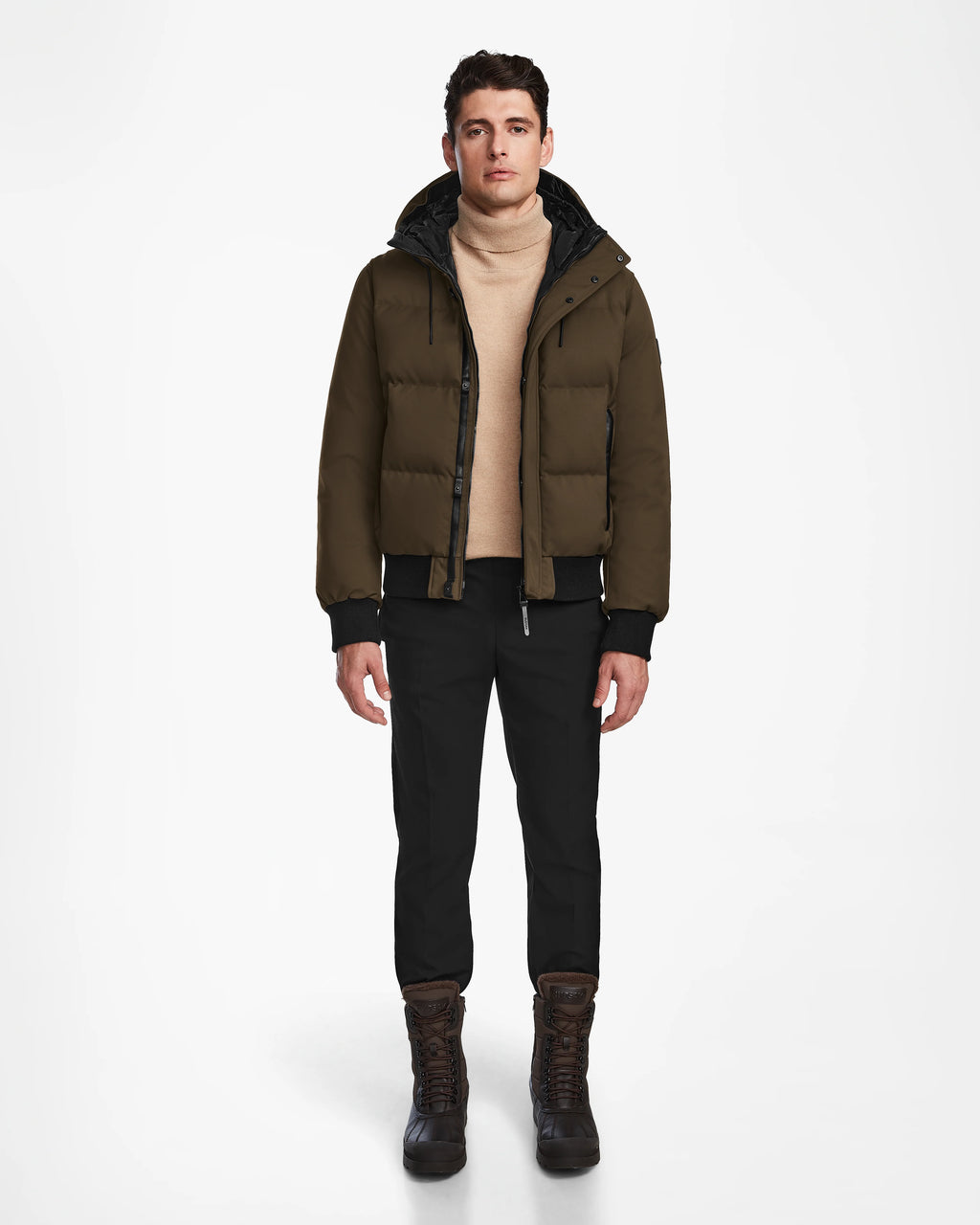 A man stands facing forward against a plain white background, wearing the Rudsak DALLAS - M122501 Army weather-resistant puffer jacket, a beige turtleneck sweater, black pants, and brown winter boots.