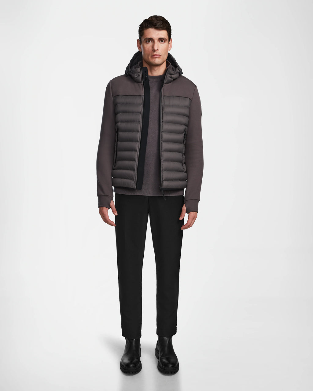 A man stands facing forward, wearing the Rudsak DURIN M - M123515 Grey mens hooded jacket with lightweight down insulation, paired with black pants and black Chelsea boots against a plain white background.