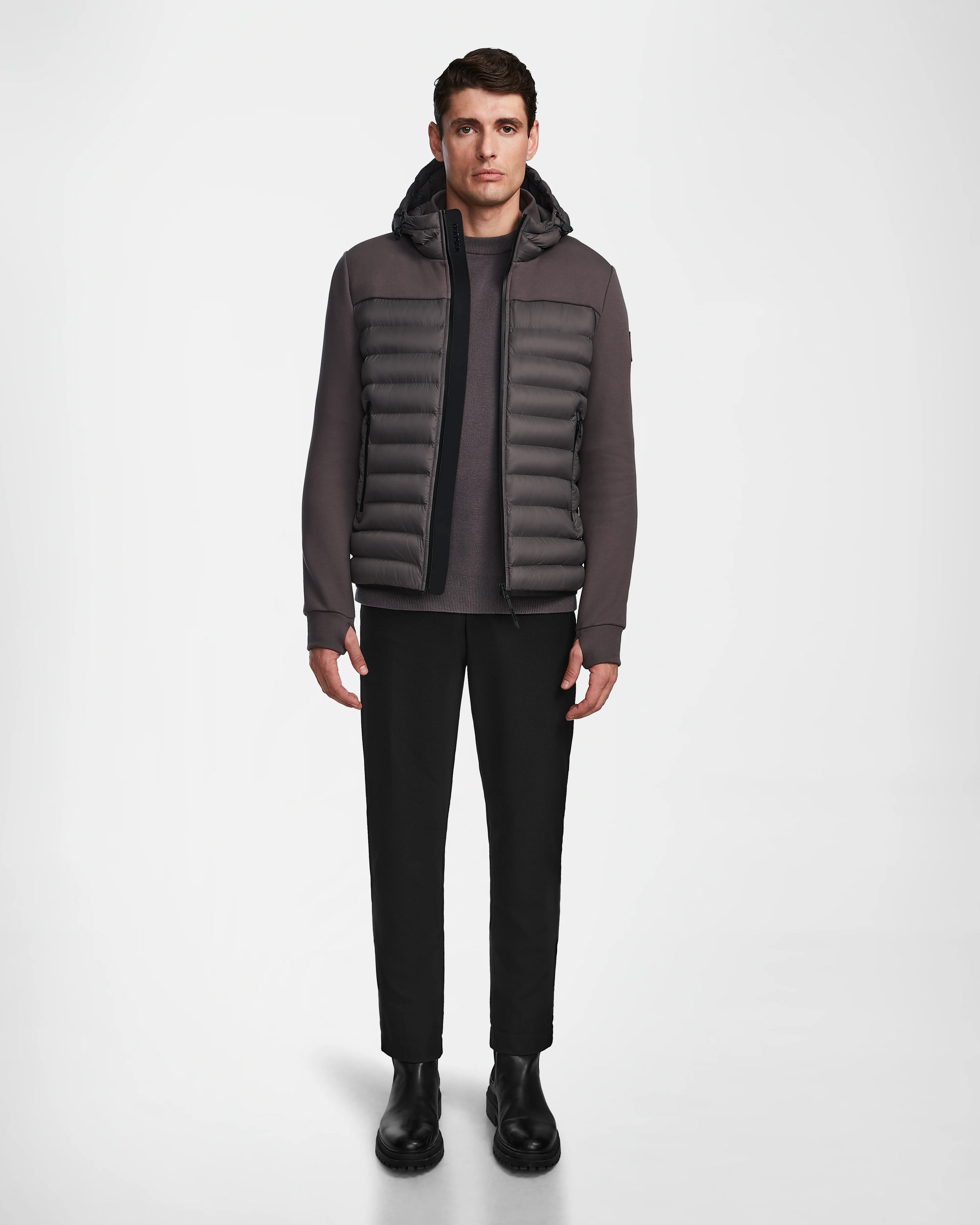 A man stands facing forward, wearing the Rudsak DURIN M - M123515 Grey mens hooded jacket with lightweight down insulation, paired with black pants and black Chelsea boots against a plain white background.