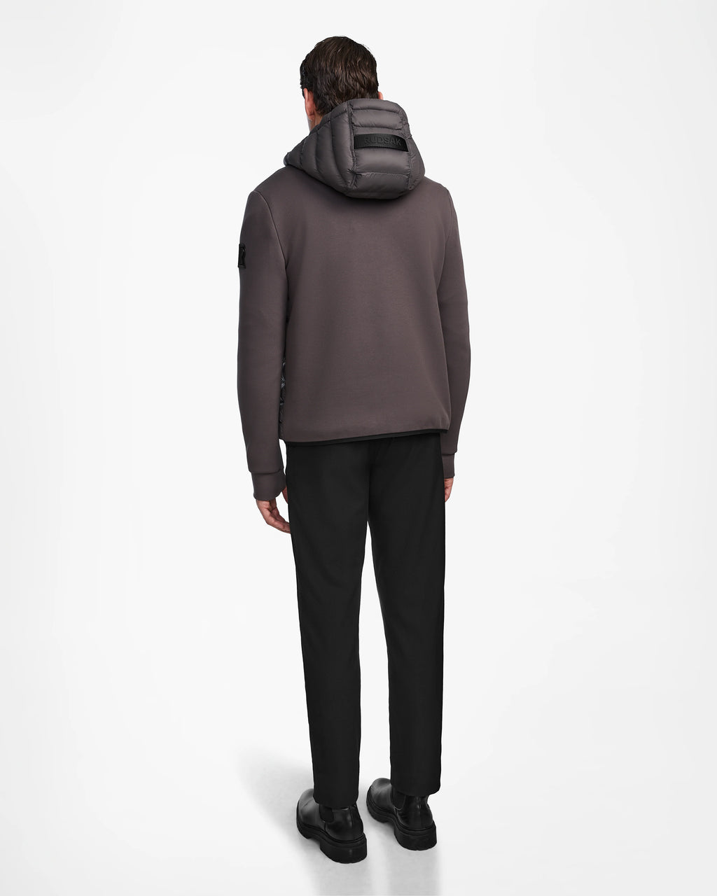 A man, facing away, wears the Rudsak DURIN M - M123515 Grey mens hooded jacket with waterproof zippers, black pants, and black shoes against a plain white background.