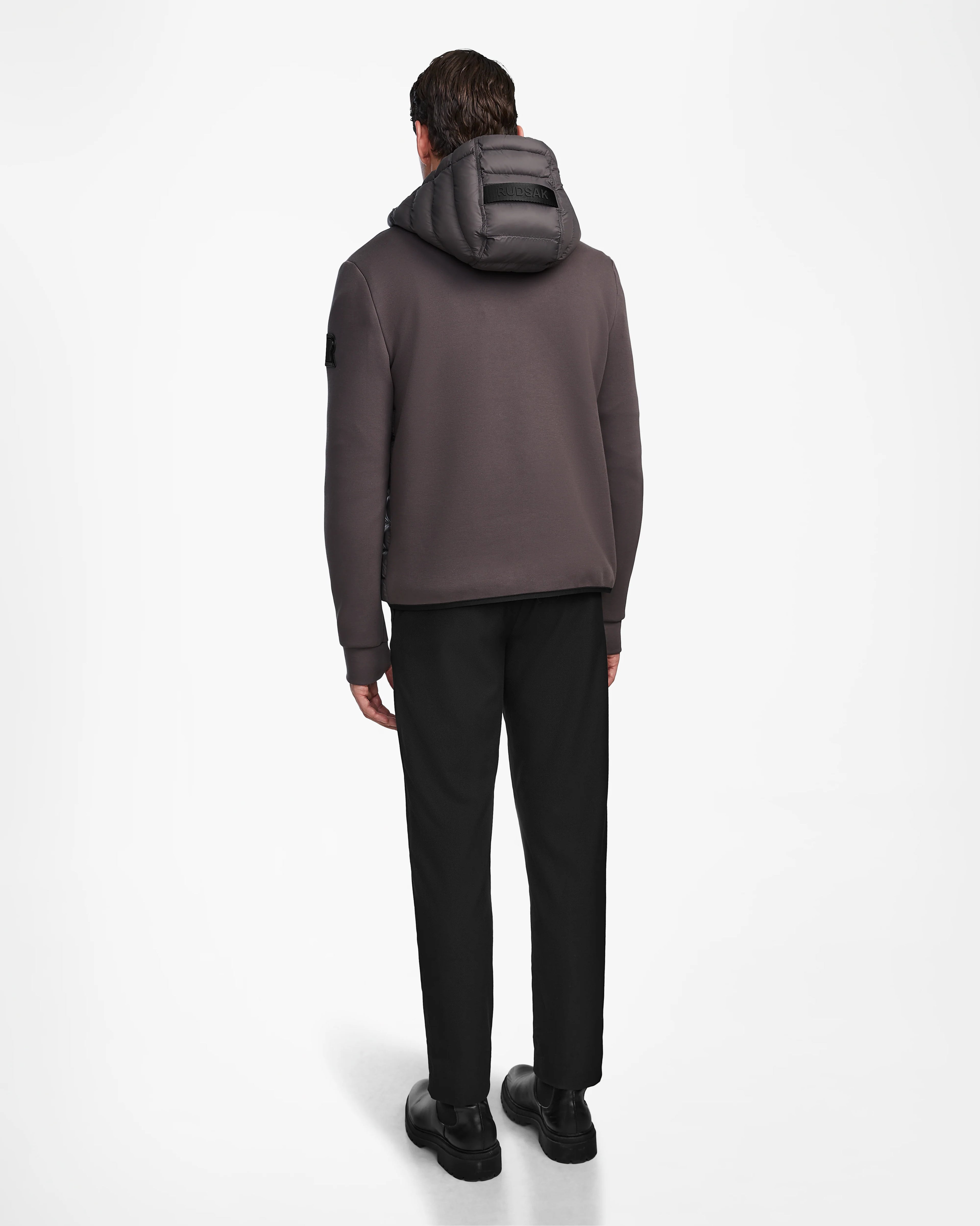 A man, facing away, wears the Rudsak DURIN M - M123515 Grey mens hooded jacket with waterproof zippers, black pants, and black shoes against a plain white background.