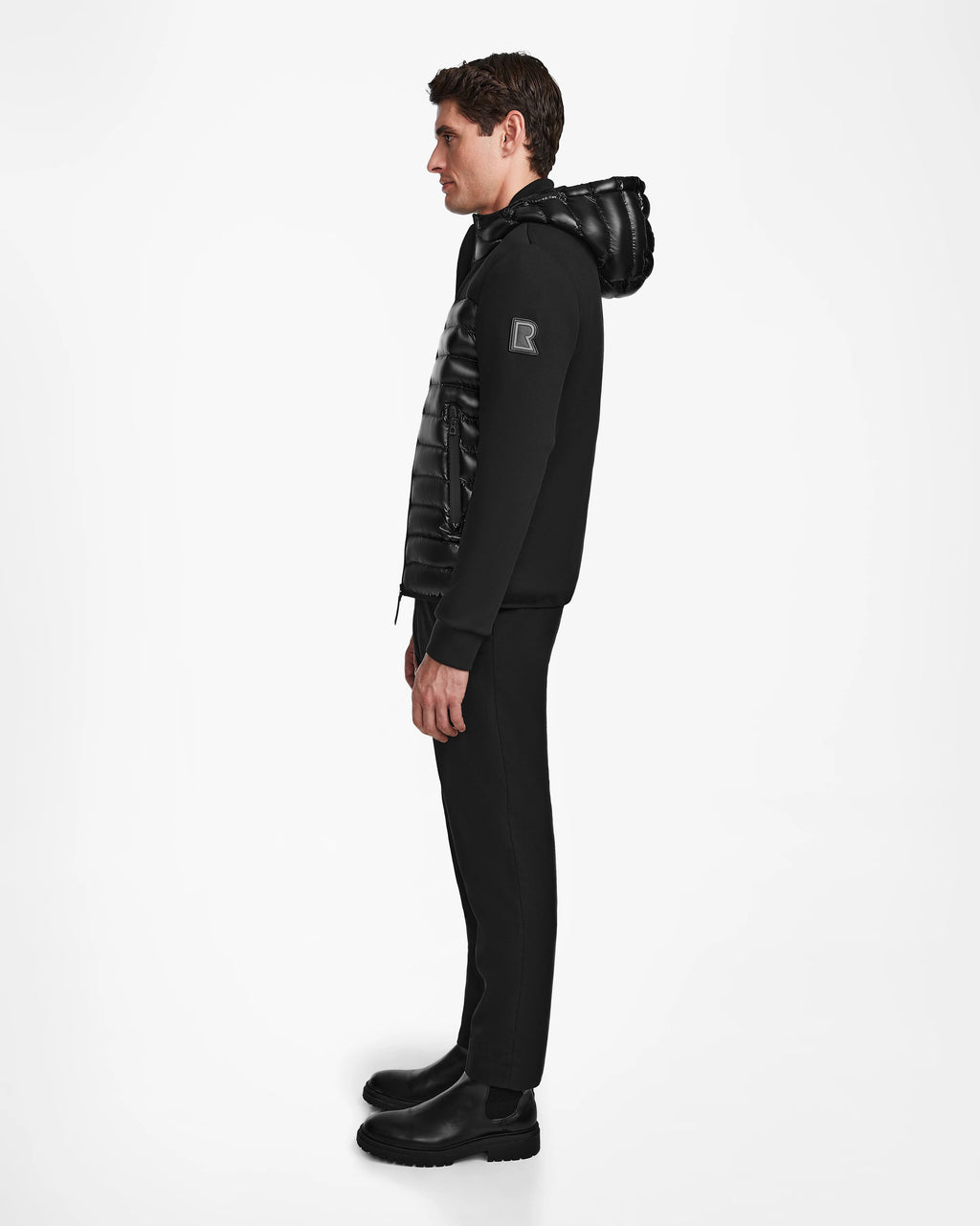 A man in profile faces left, wearing the Rudsak DURIN S - M123518 Black mens hooded jacket with lightweight down insulation, paired with matching black pants and shoes against a plain white background.