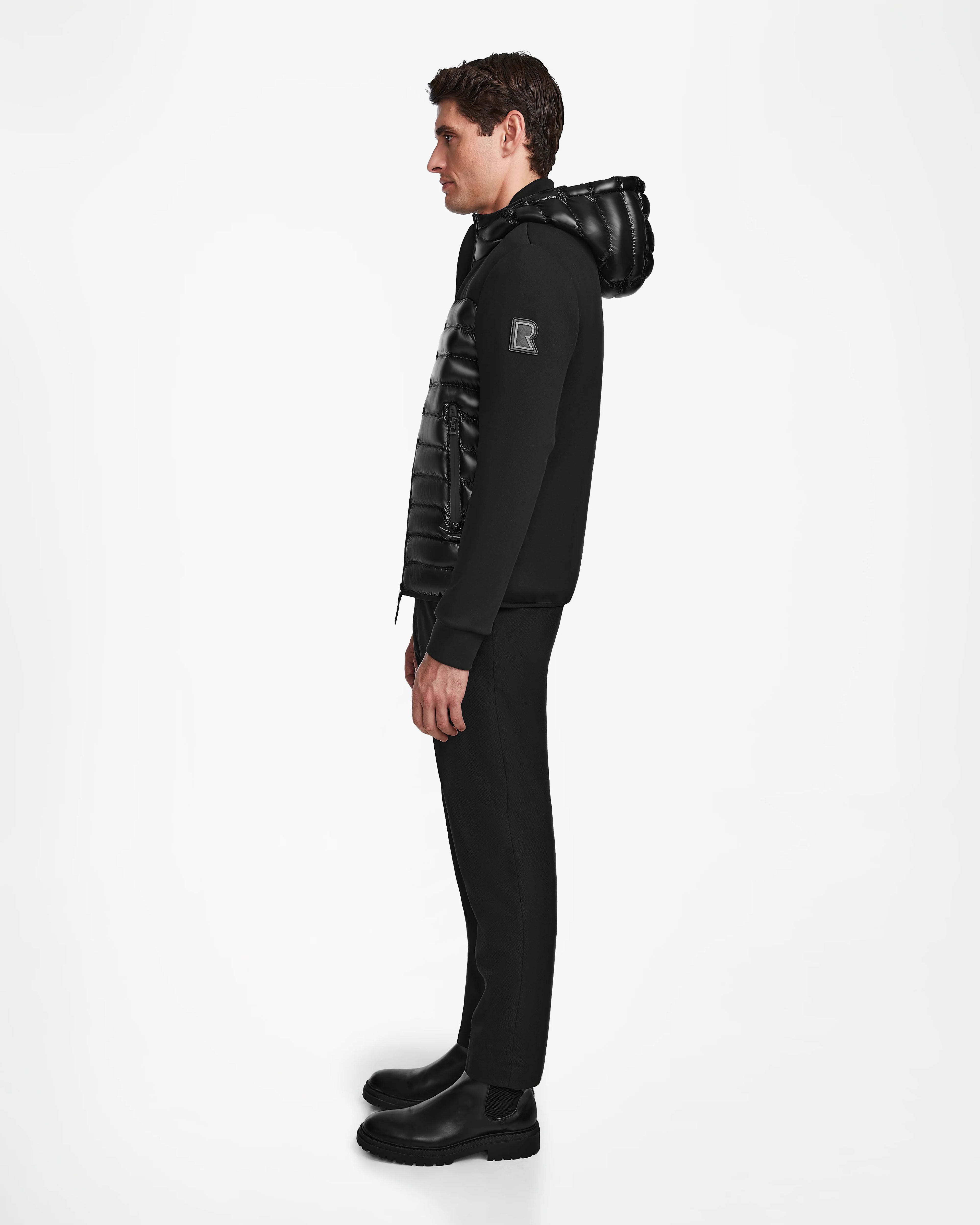 A man in profile faces left, wearing the Rudsak DURIN S - M123518 Black mens hooded jacket with lightweight down insulation, paired with matching black pants and shoes against a plain white background.