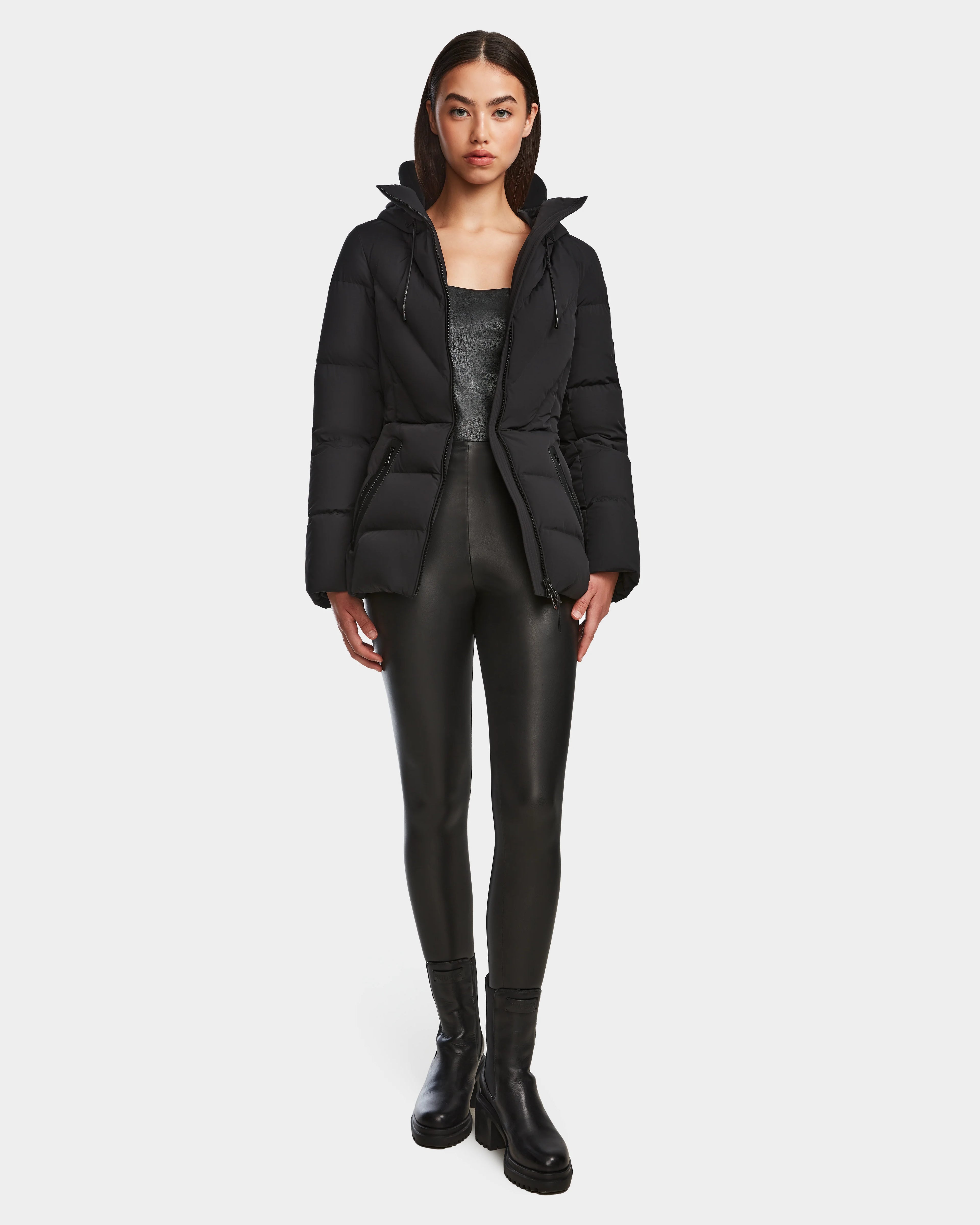 A woman faces forward against a white background, wearing the Rudsak ENZAH - 8124692 Black mid-length puffer jacket, a dark top, black faux leather leggings, and black ankle boots.