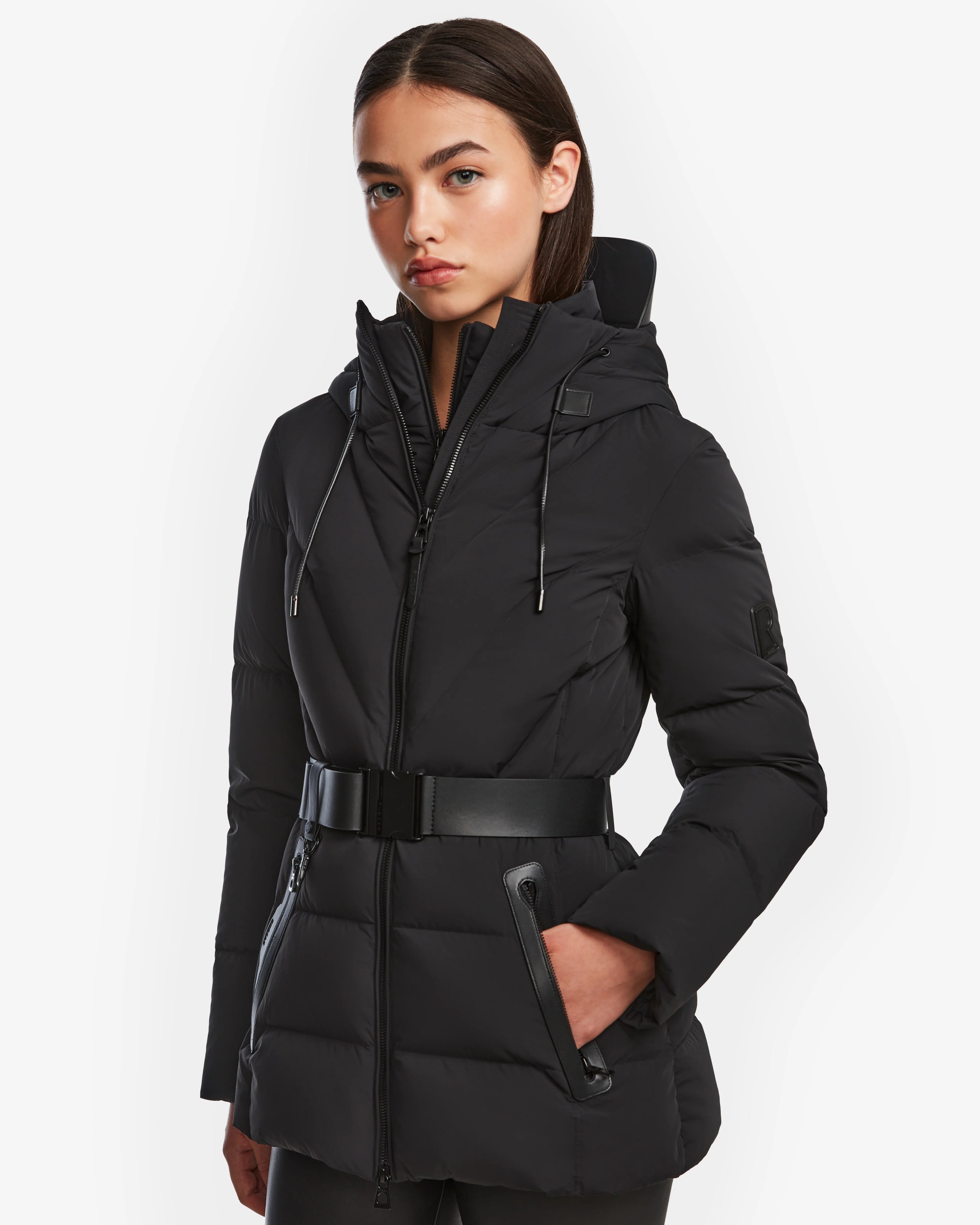 A woman wears the Rudsak ENZAH - 8124692 Black mid-length puffer jacket with a hood, standing forward with one hand in her pocket against a white background. This cozy down coat is perfect for winter.