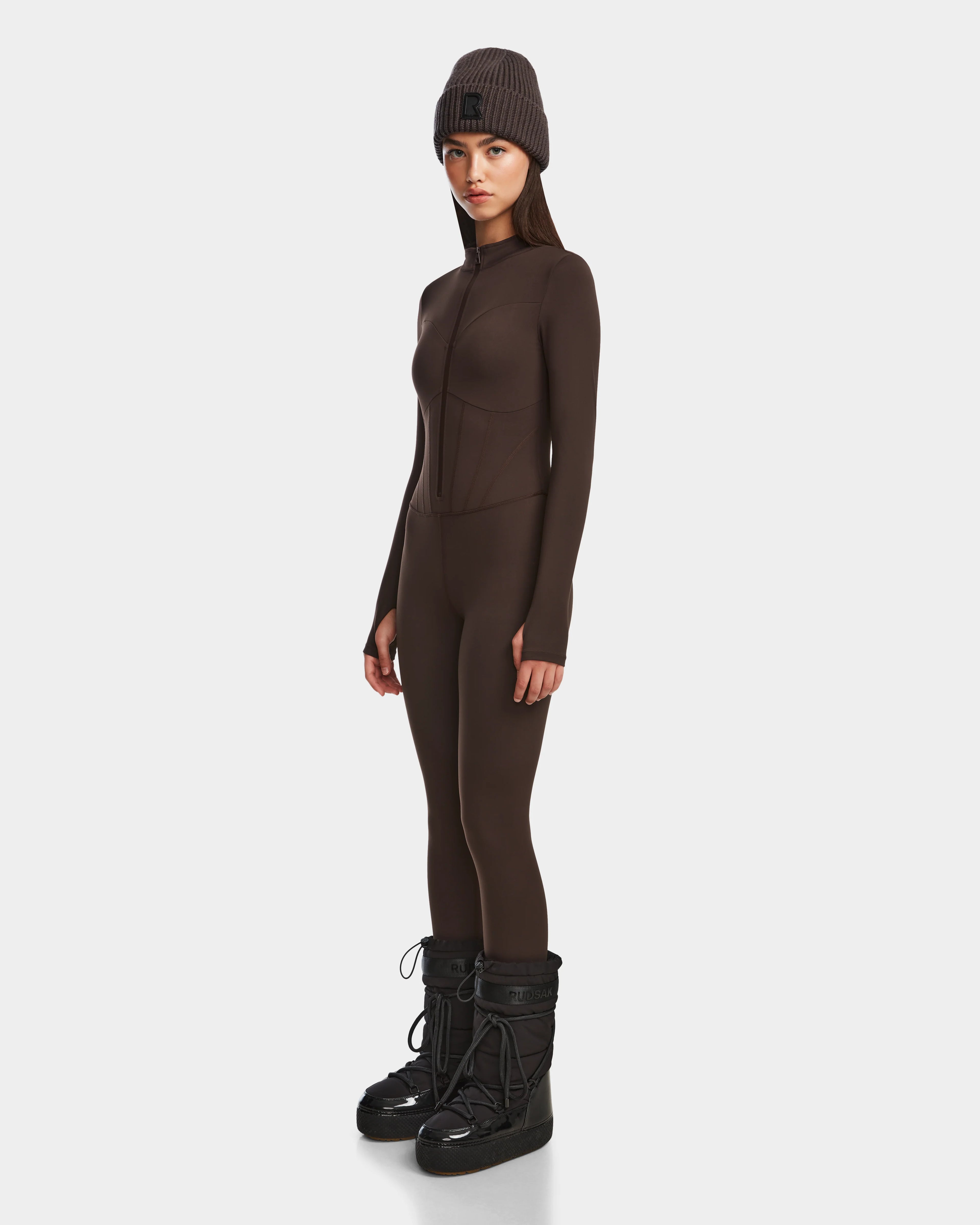 A woman faces slightly sideways, wearing the Rudsak IVY - 8524599 Espresso fitted women’s stretch bodysuit jumpsuit with a matching beanie and black winter boots, creating a ski-chic look against a plain white background.
