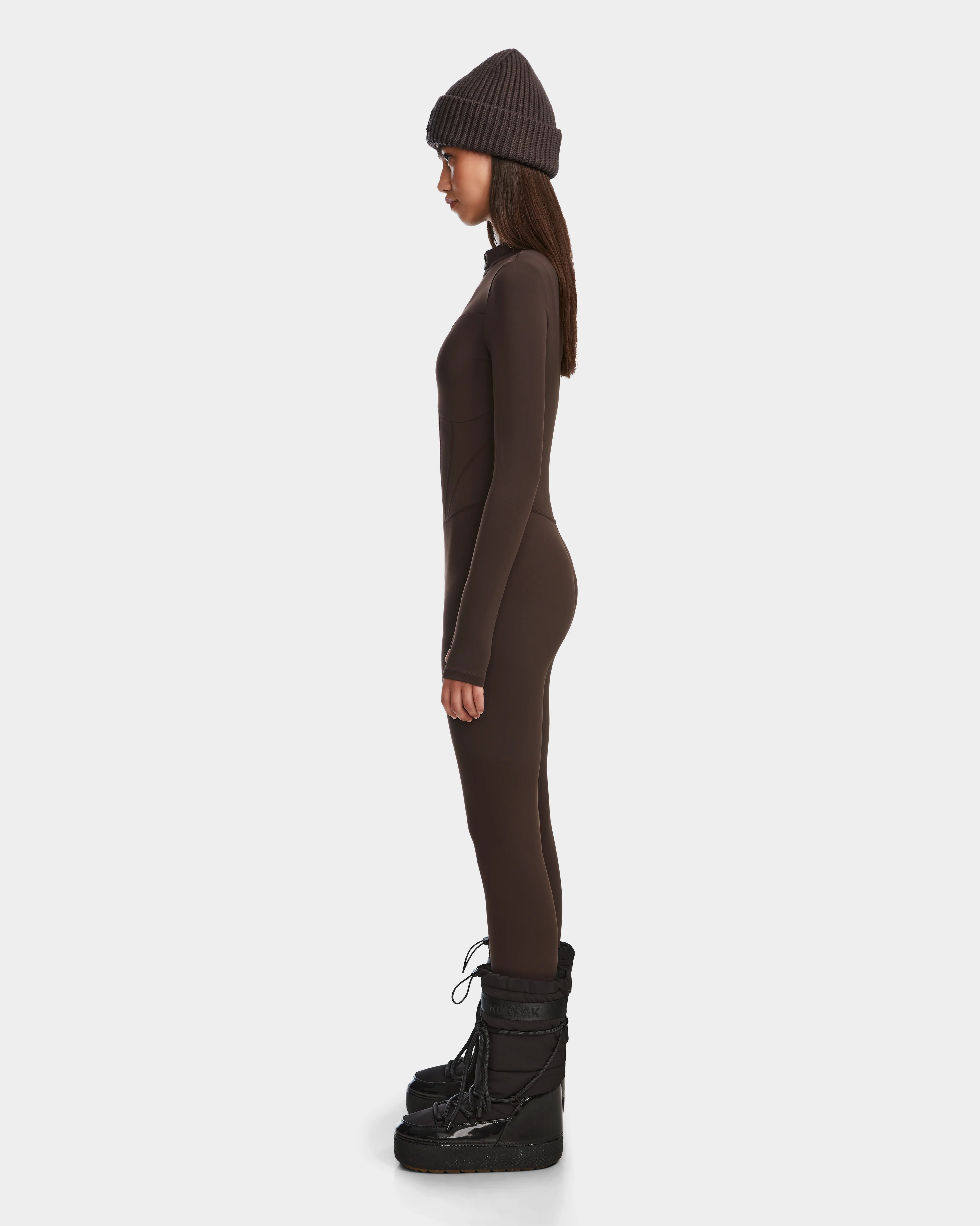 A woman stands in profile wearing the Rudsak IVY - 8524599 Espresso stretch bodysuit with a zip-up front, paired with a matching knit beanie and black winter boots, showcasing ski-chic style against a plain white background.