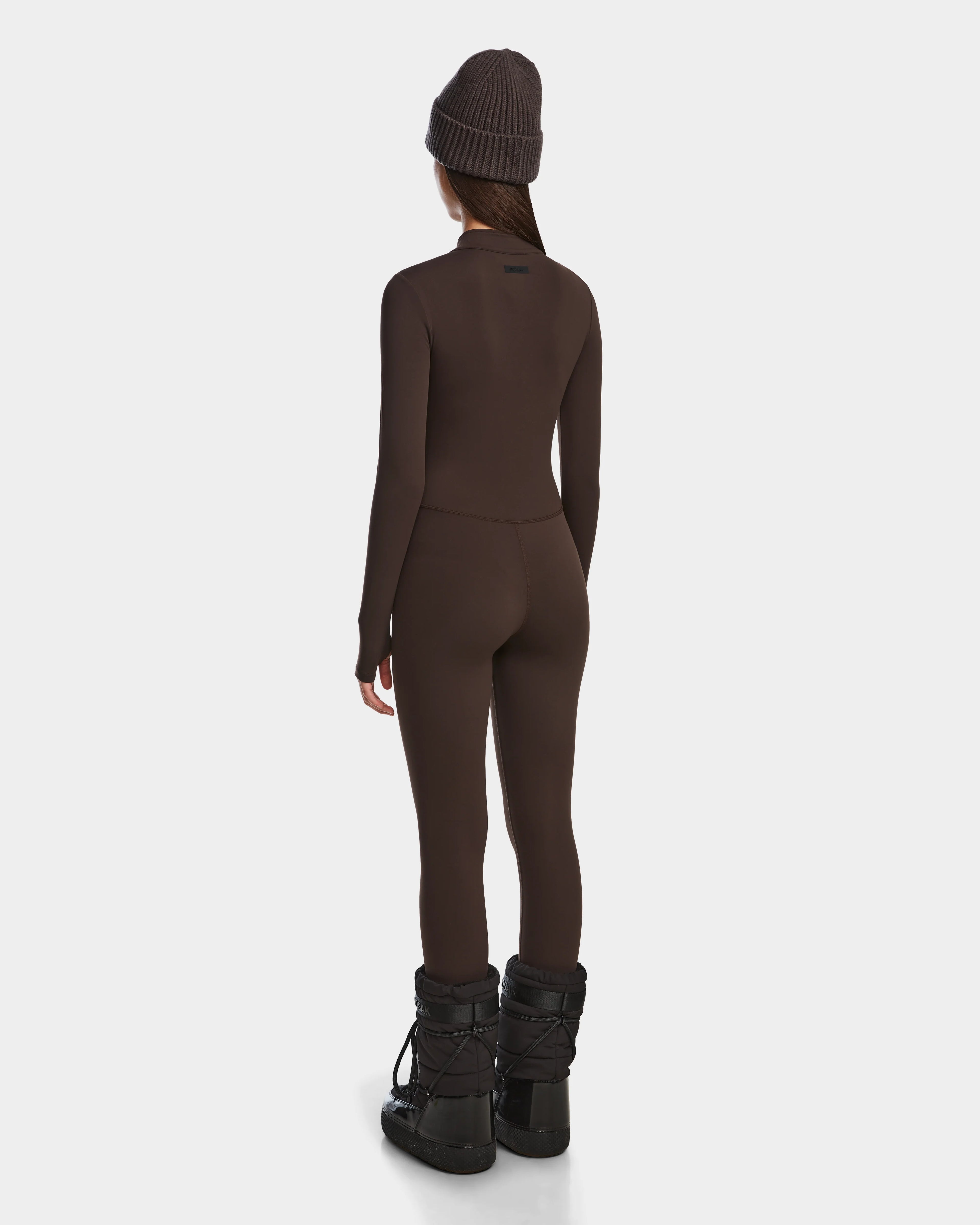 A woman faces away, wearing the Rudsak IVY - 8524599 Espresso fitted stretch bodysuit with a matching beanie and black winter boots, highlighting ski-chic style against a plain white backdrop.