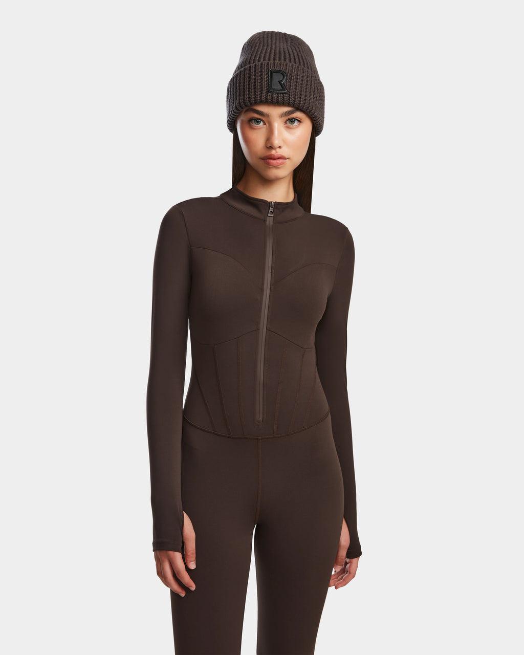 A woman in the Rudsak IVY - 8524599 Espresso fitted, long-sleeve stretch bodysuit with a zip-up front and a ribbed dark gray beanie stands against a white background, looking forward with a neutral expression.