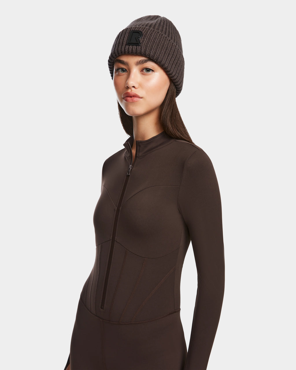 A woman wears the Rudsak IVY - 8524599 Espresso fitted long-sleeve zip-up bodysuit and a ribbed gray beanie, standing against a plain white background with a neutral expression for a ski-chic look.
