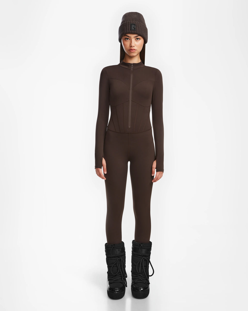 A person stands against a white background wearing the Rudsak IVY - 8524599 Espresso fitted women’s stretch bodysuit jumpsuit with a matching brown beanie and black snow boots, hands at their sides and facing forward with a neutral expression.