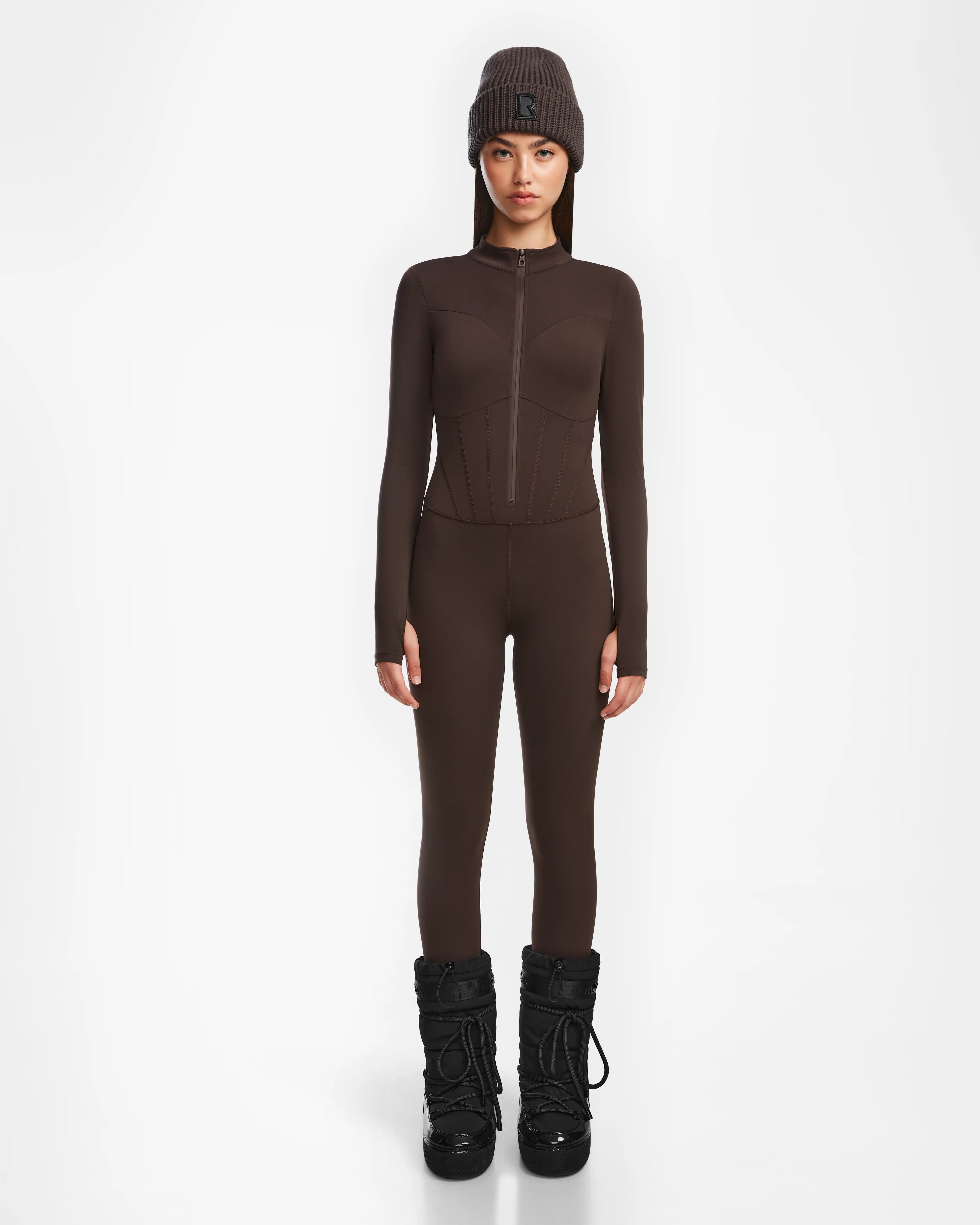 A person stands against a white background wearing the Rudsak IVY - 8524599 Espresso fitted women’s stretch bodysuit jumpsuit with a matching brown beanie and black snow boots, hands at their sides and facing forward with a neutral expression.