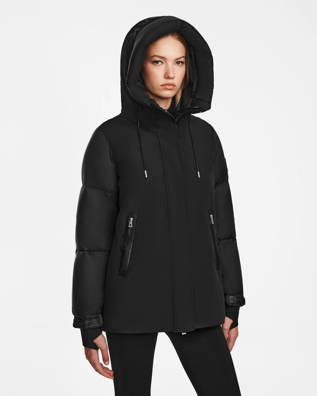 A woman stands against a plain white background, wearing the Rudsak LOLI - 8124507 Black womens down parka with a hood and zippered pockets, looking forward with a neutral expression.