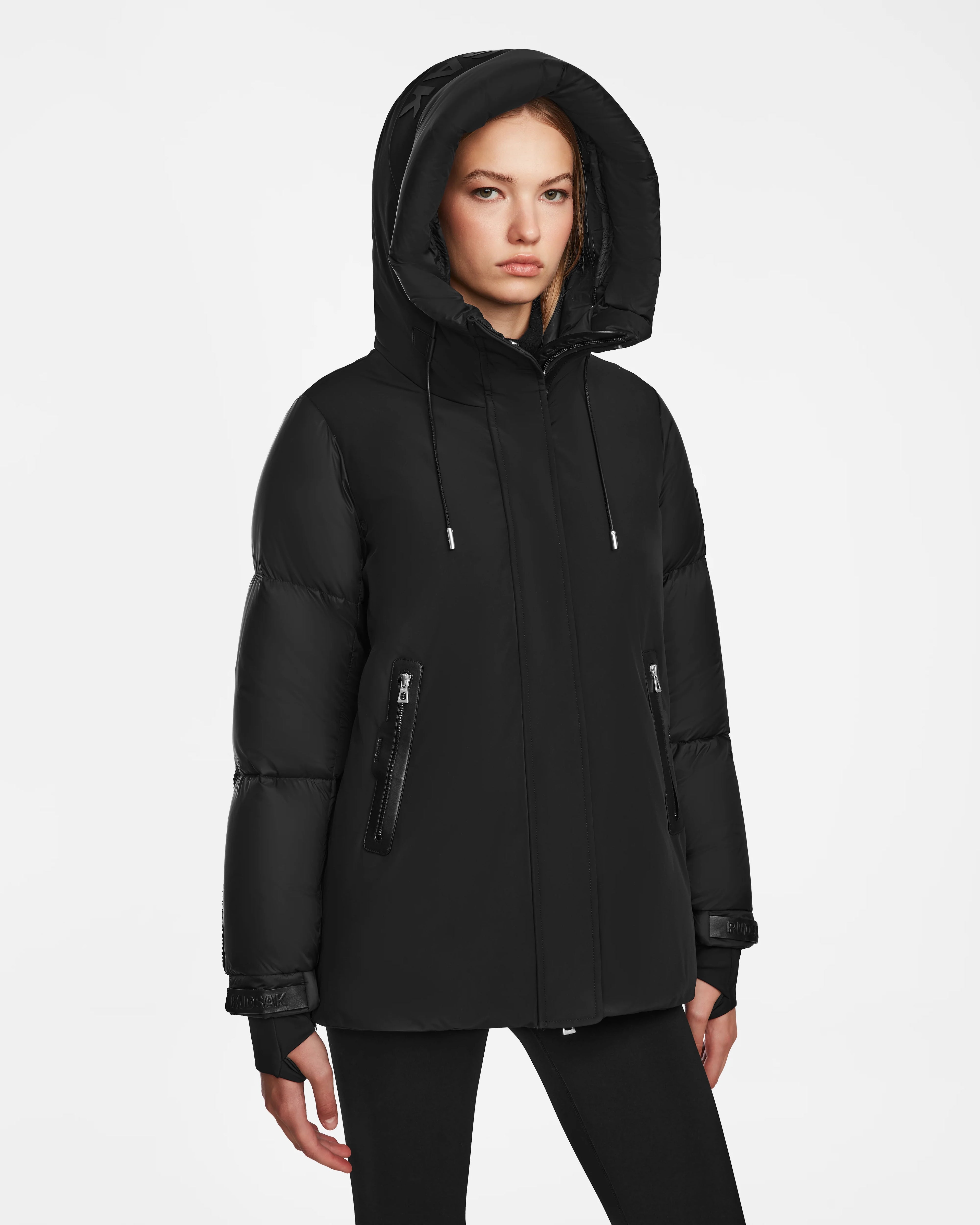 A woman stands against a plain white background, wearing the Rudsak LOLI - 8124507 Black womens down parka with a hood and zippered pockets, looking forward with a neutral expression.